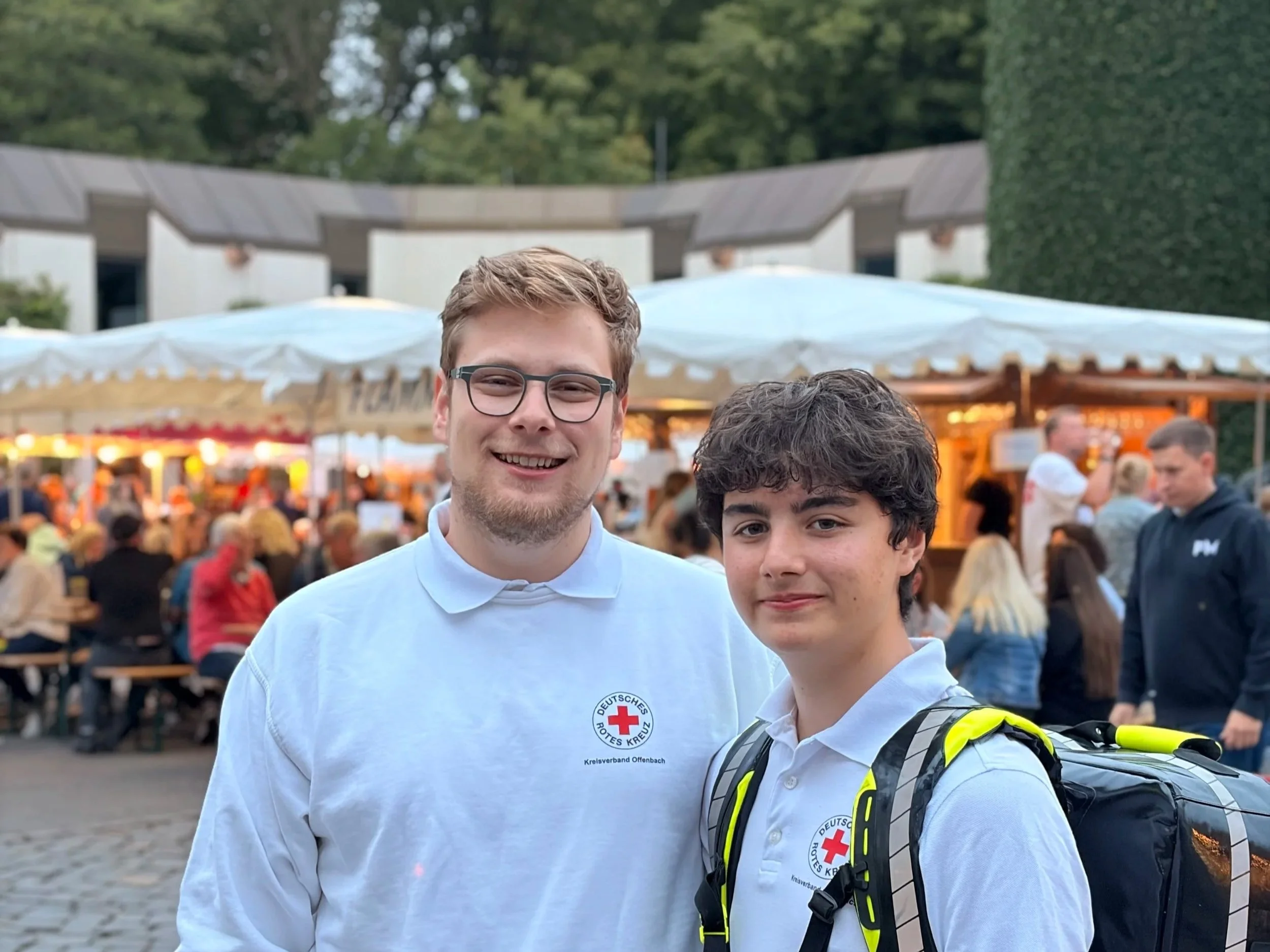 Zwei junge Männer, einer trägt eine Jacke mit dem Logo der Deutschen Rotes Kreuz, beim öffentlichen Event im Freien, im Hintergrund ein Markt mit Ständen und vielen Menschen.
