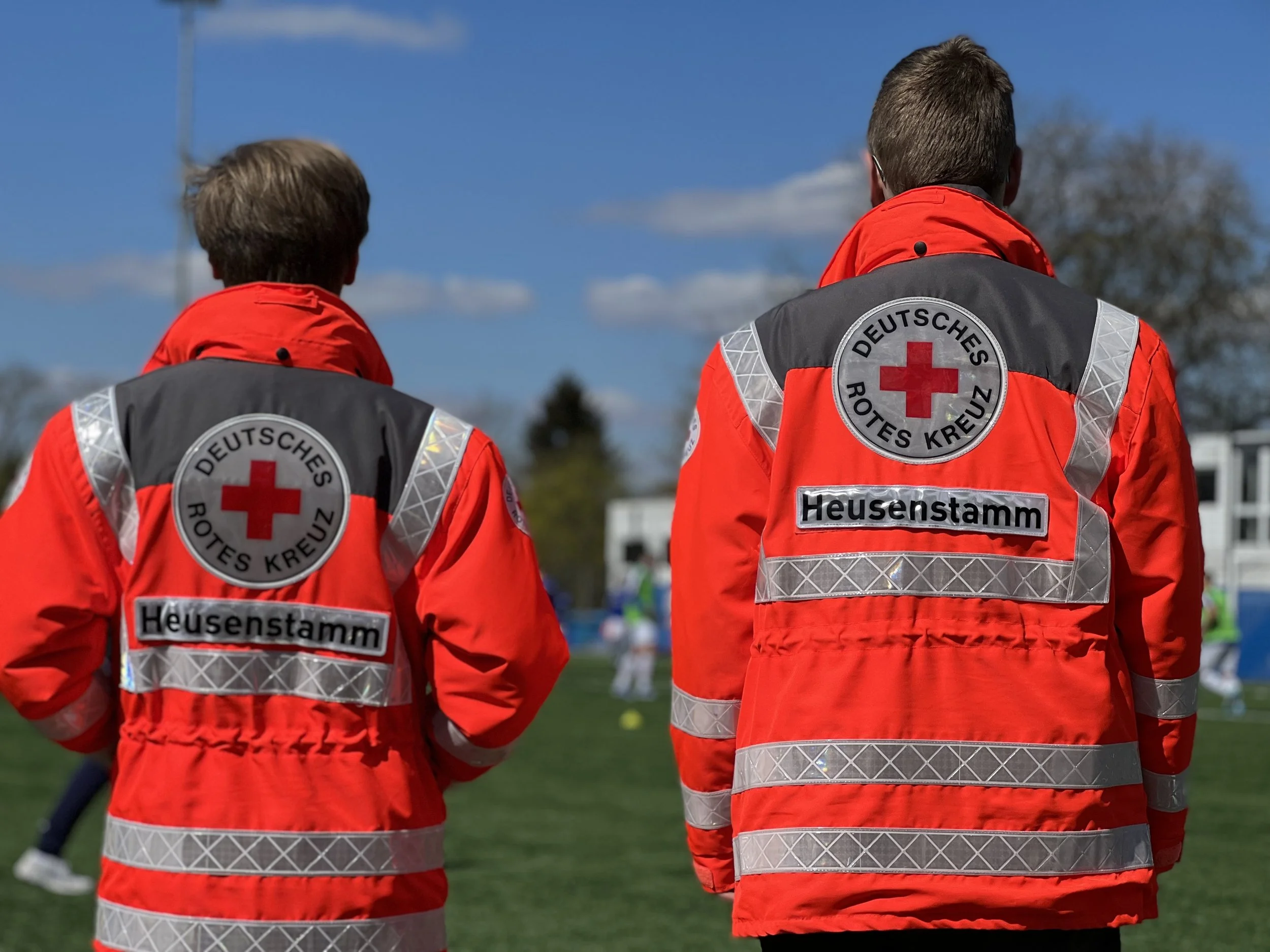 Zwei Personen von hinten in roten Jacken mit Deutsche Rotes Kreuz-Emblemen, am Rand eines Sportplatzes.