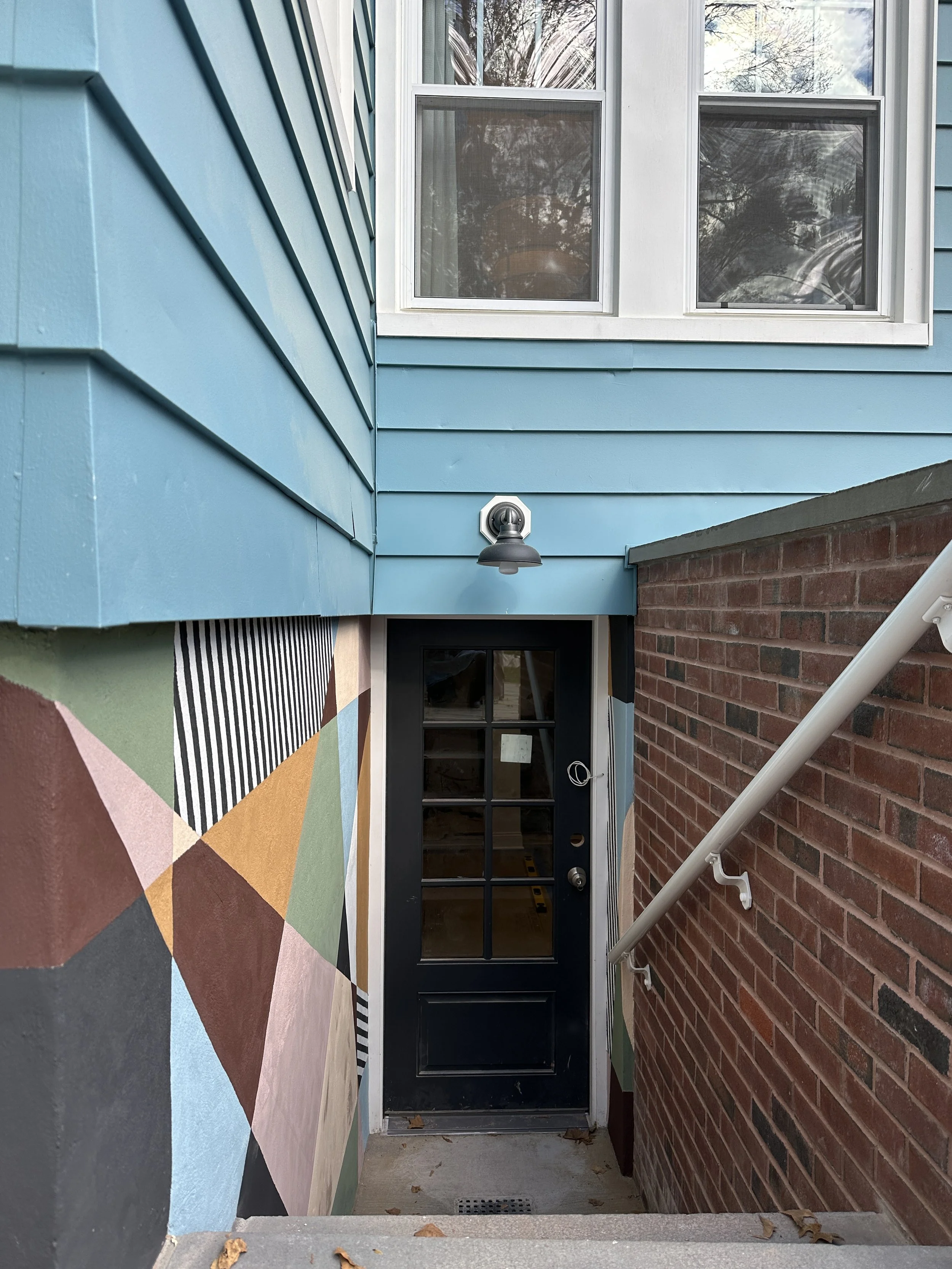 Custom basement entryway mural by Shapes For The People, painted with bold geometric shapes and earthy tones along an exterior stairwell.