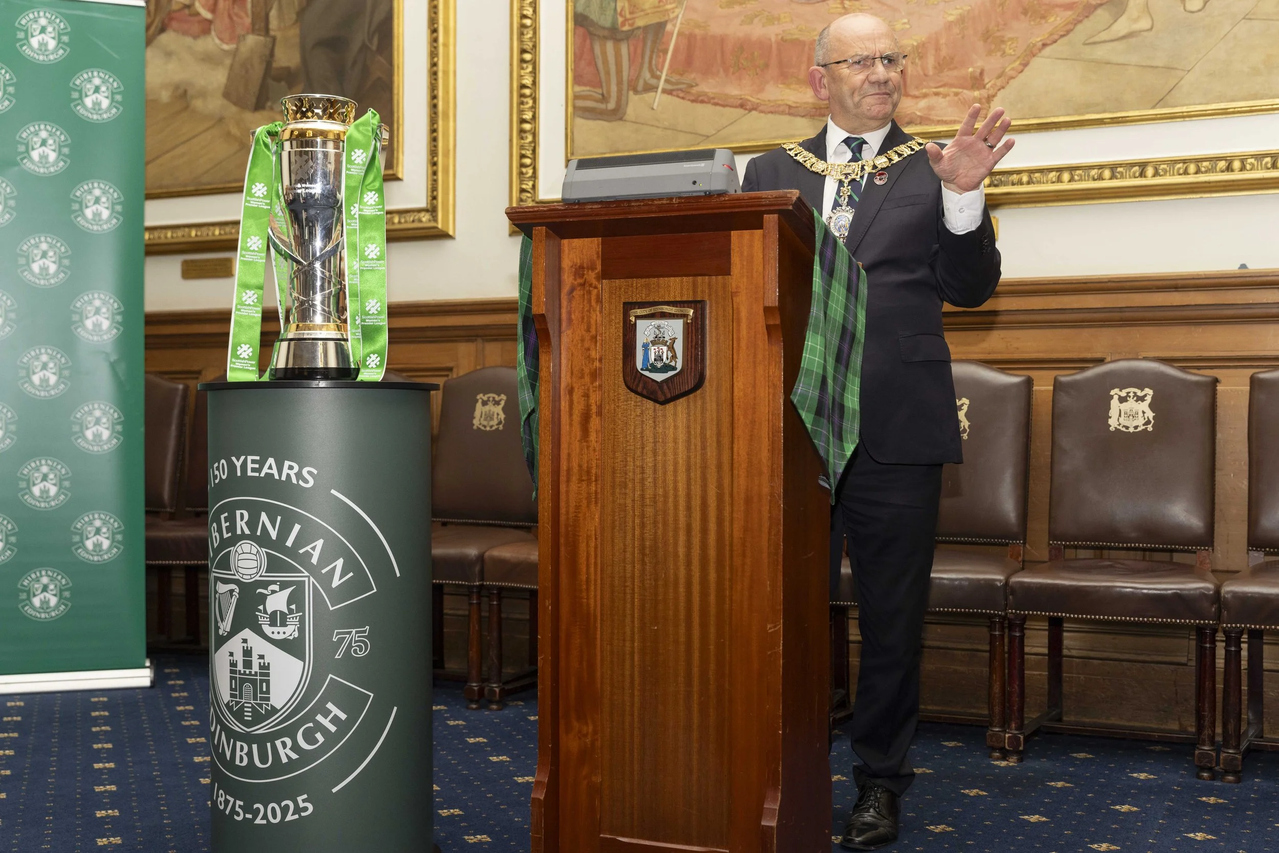 A man in formal attire, wearing a tartan sash and gold chain, stands at a wooden podium in a decorated room with wood-paneled walls and framed artwork. To his left, there is a green stand with a trophy and a banner celebrating 150 years of the Aberlour Scottish whisky from 1875 to 2025.