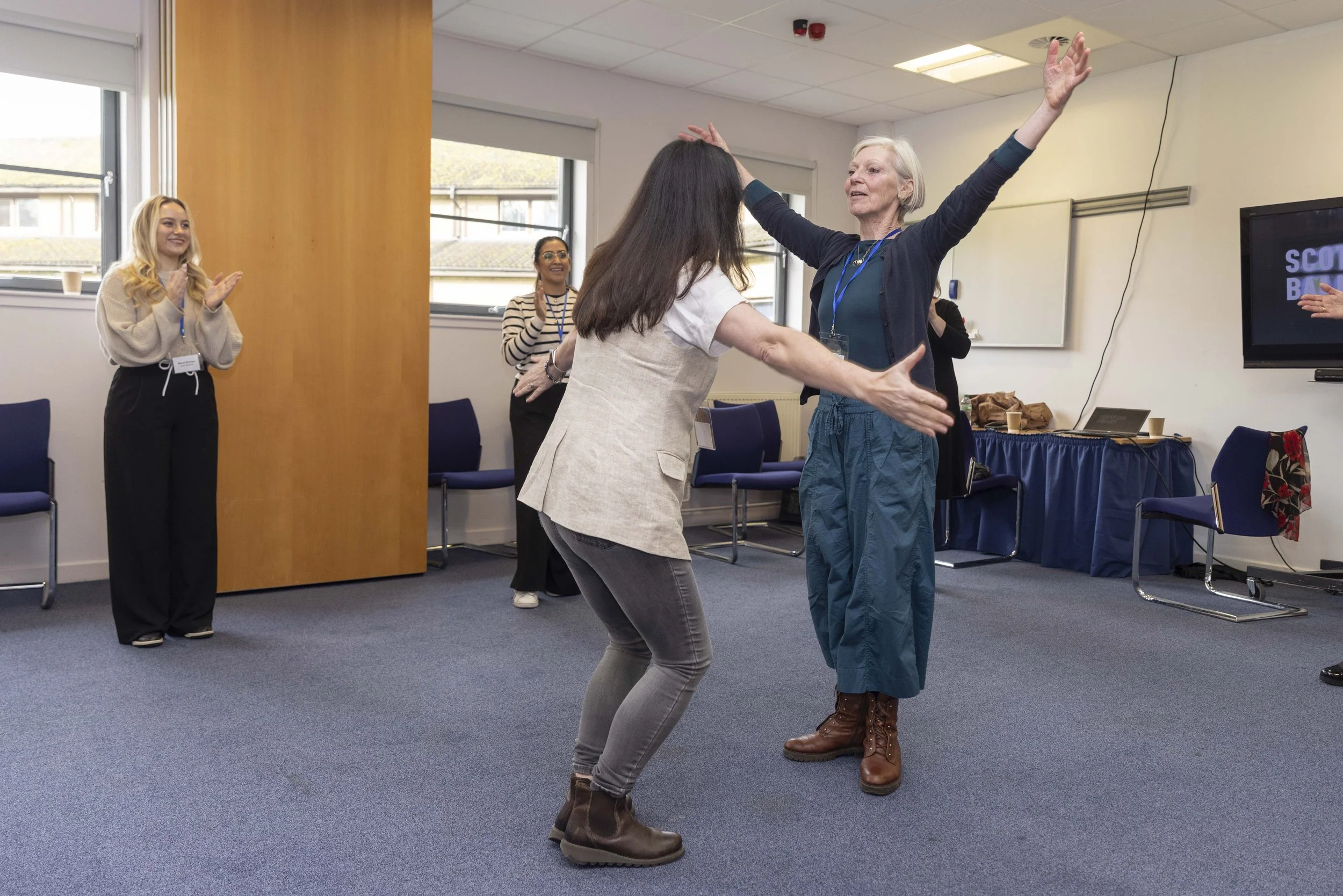 Two women are dancing in a room with three women watching and clapping in the background.