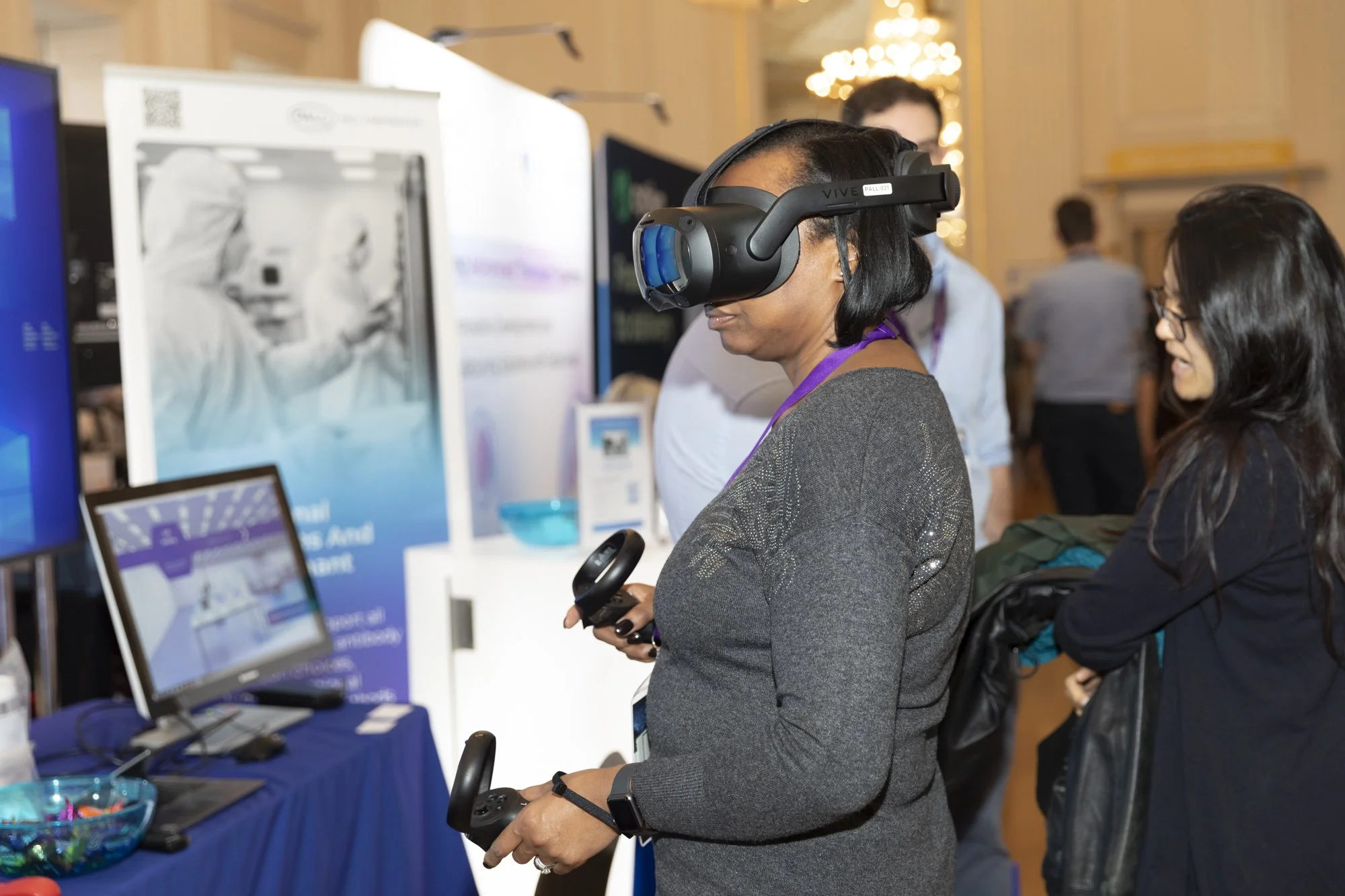A woman wearing a VR headset and holding a gaming controller in a convention setting, with other attendees and display screens nearby.