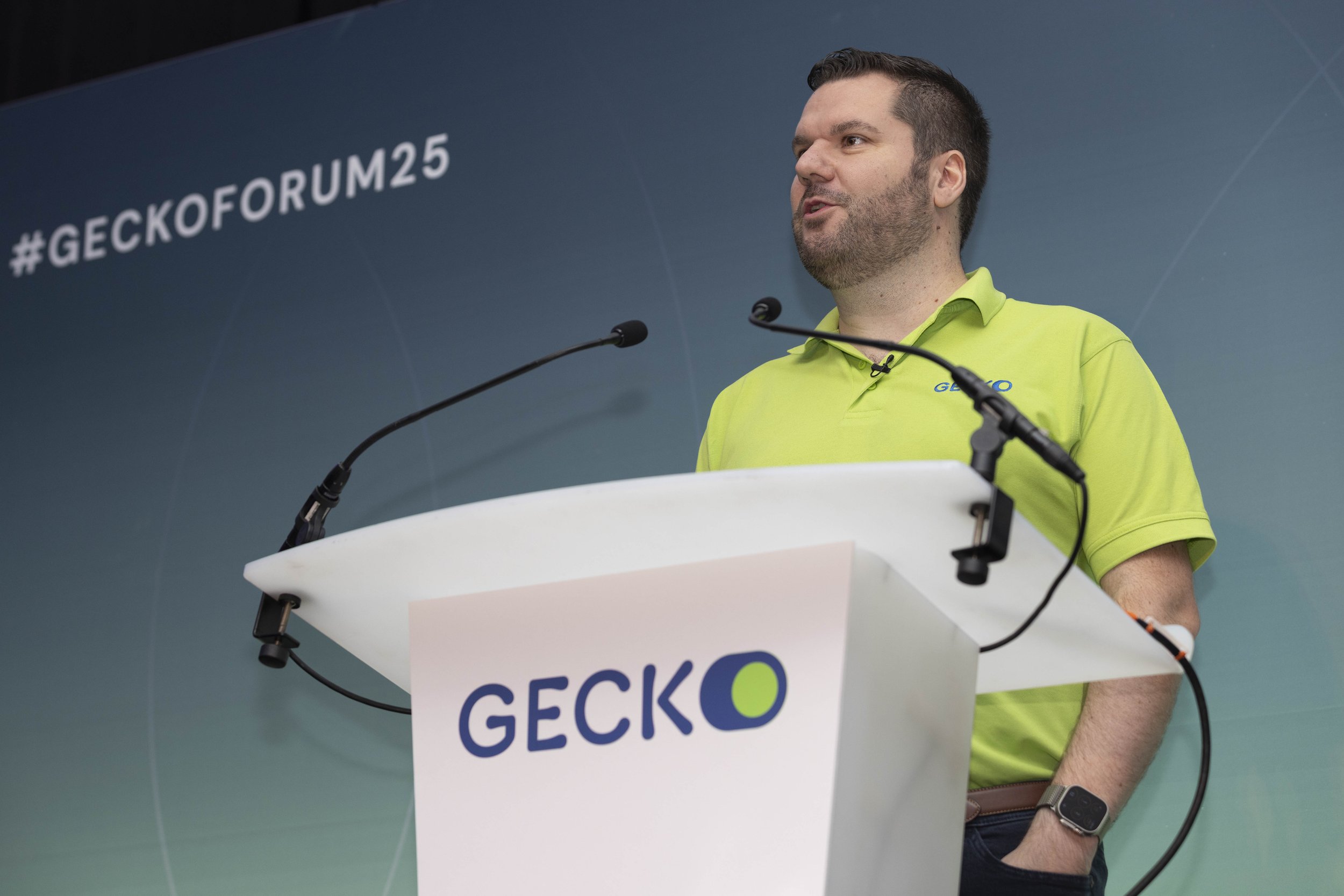 Man in lime green polo shirt giving speech at Geico conference with hashtag #GECKOFORUM25 on blue-green backdrop.