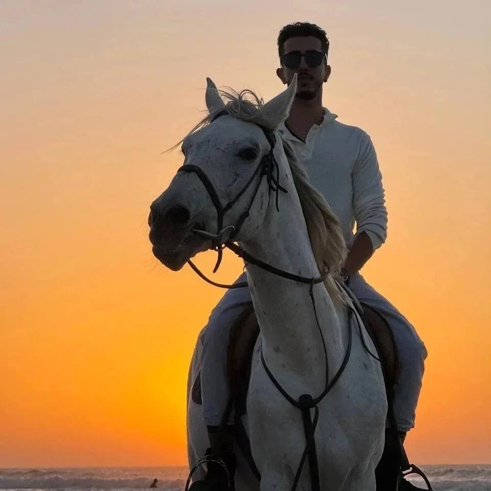 A man wearing sunglasses and a white shirt is riding a white horse against a sunset background.