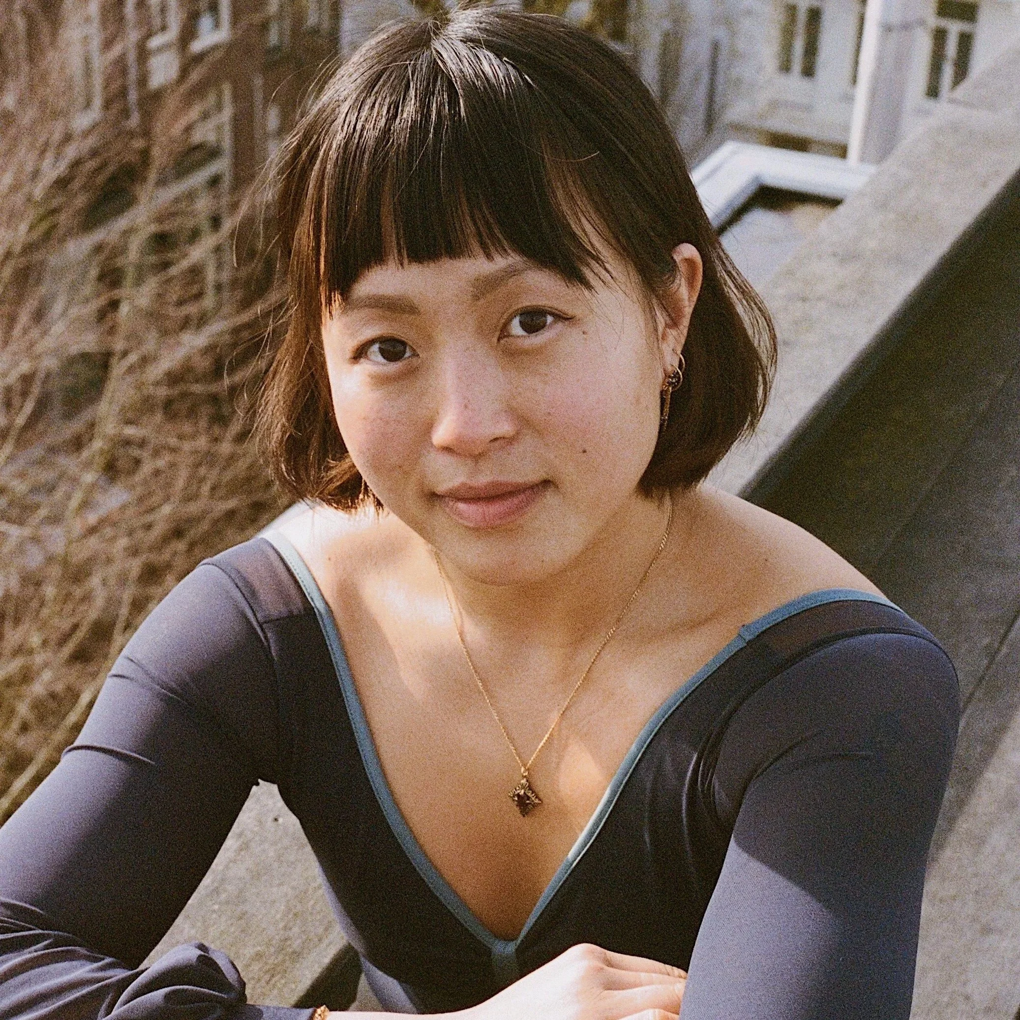 A young woman with short brown hair, wearing a black long-sleeve shirt with blue trim, a gold necklace, and earrings. She is sitting outdoors near a wrought iron fence, with a concrete surface and building visible in the background.