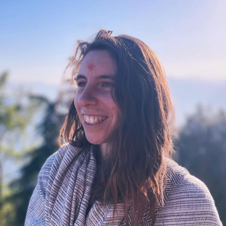 A woman with long brown hair, a nose piercing, and a small red mark on her forehead, smiling outdoors during the daytime, with trees and a blue sky in the background.