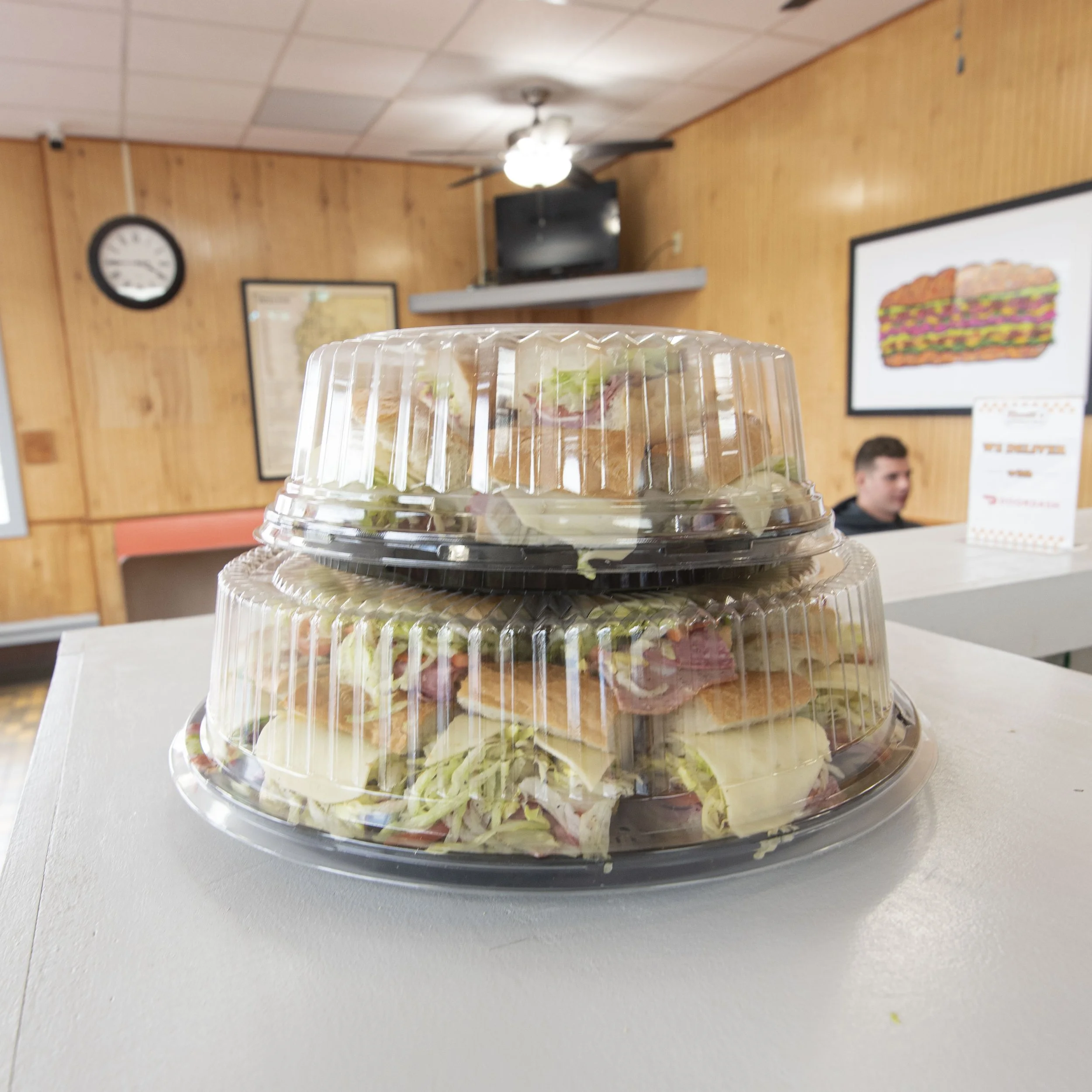 Two stacked plastic containers filled with sandwiches containing lettuce, tomato, cheese, and meat, placed on a white table inside a restaurant with wooden walls, a clock, and a television in the background.