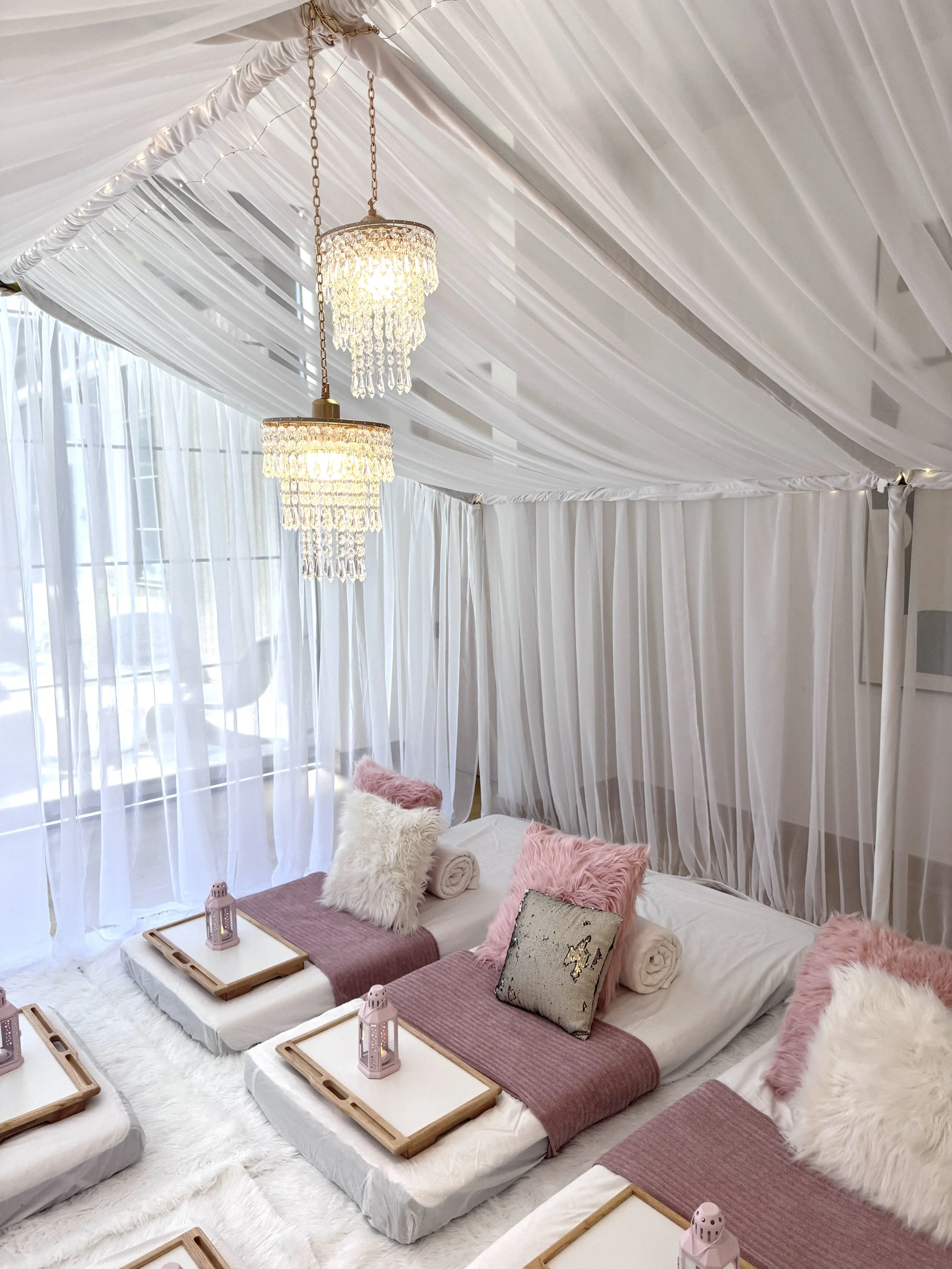 A cozy indoor setting with a white canopy tent, decorated with white drapes and string lights hanging from the ceiling. There are three low beds on the floor, each adorned with pink and white fluffy pillows, rolled towels, and pink mats. Small wooden trays with pink lanterns are placed on the beds.