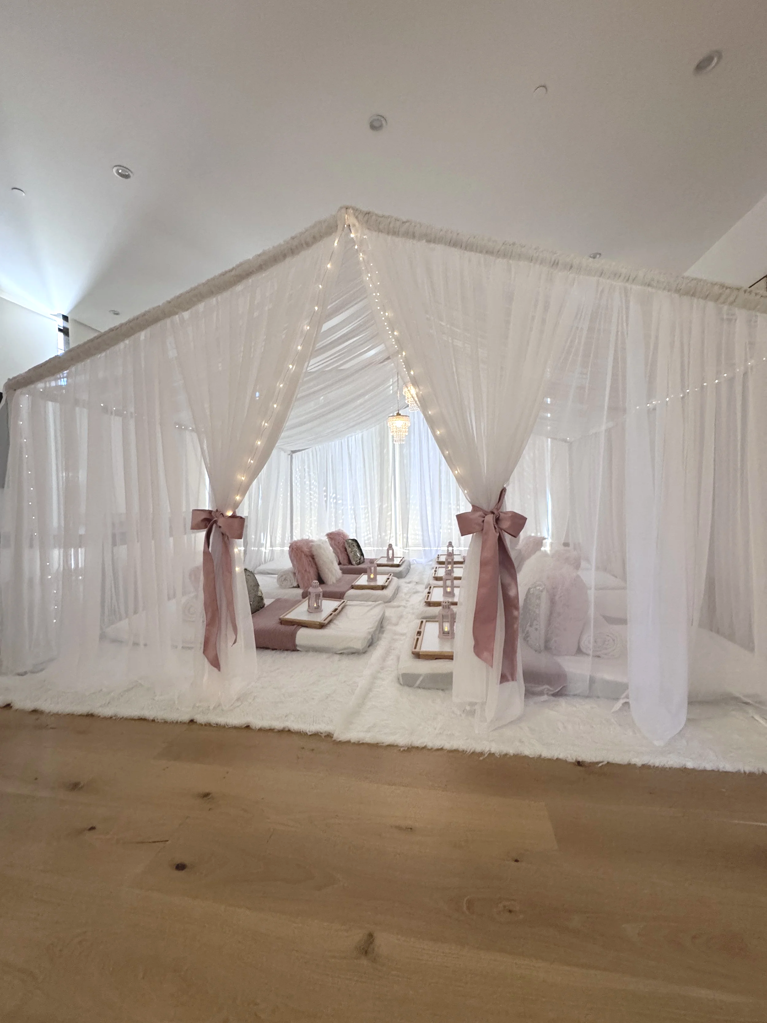 A decorated indoor space with sheer white curtains forming a canopy, soft pink ribbons tying the curtains, and a sitting area with plush pillows and small tables, illuminated by string lights and a chandelier.