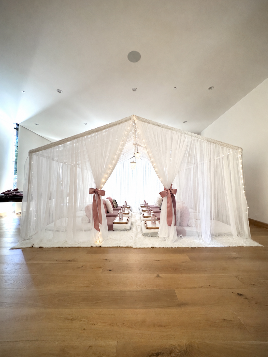 Elegant indoor sleepover with a canopy decorated with white curtains, pink ribbons, fairy lights, and plush mattresses with pink cushions.