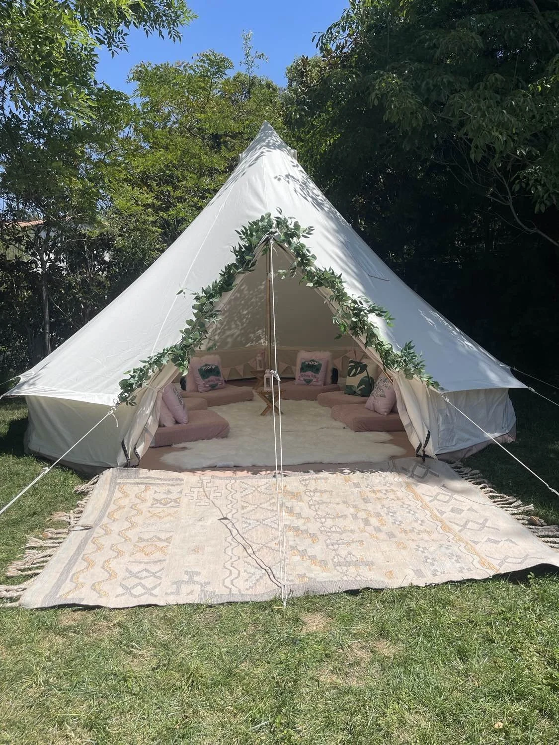 A white glampinh tent decorated with green foliage and pillows, set on a grassy lawn with a colorful patterned rug in front and surrounded by trees under a clear blue sky.