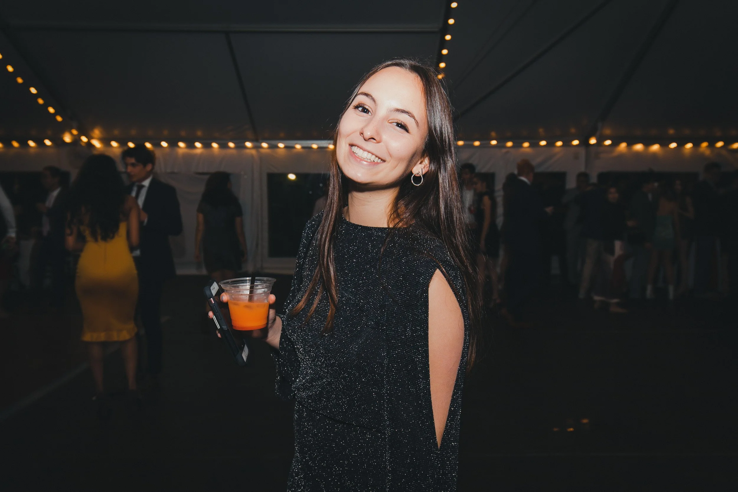 A smiling young woman with long brown hair wearing a black sparkly dress, holding a drink in a clear plastic cup, at an indoor evening event with string lights and other people in the background.