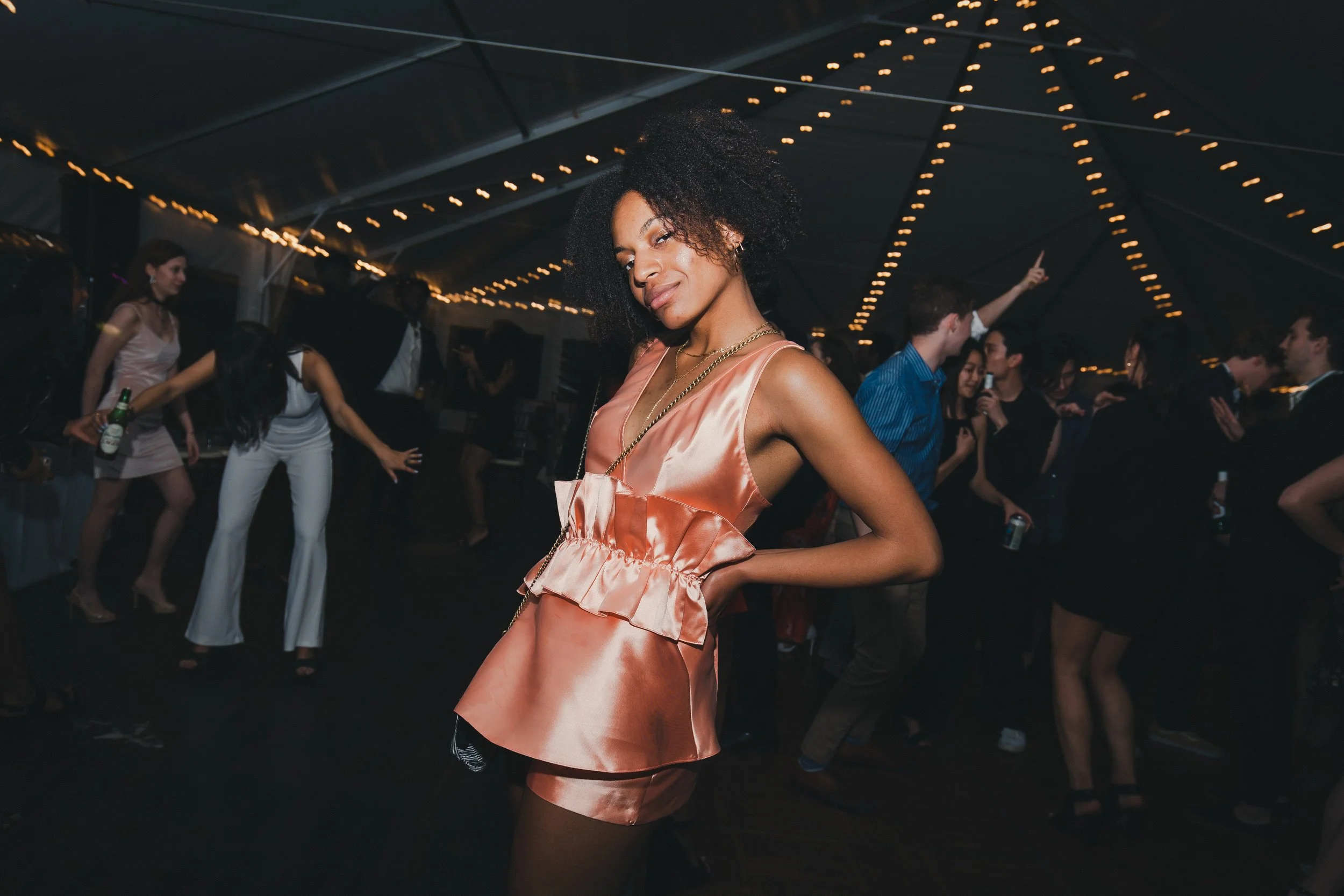 A woman in a peach satin dress posing on a dance floor at a party or celebration under string lights, with other people dancing in the background.