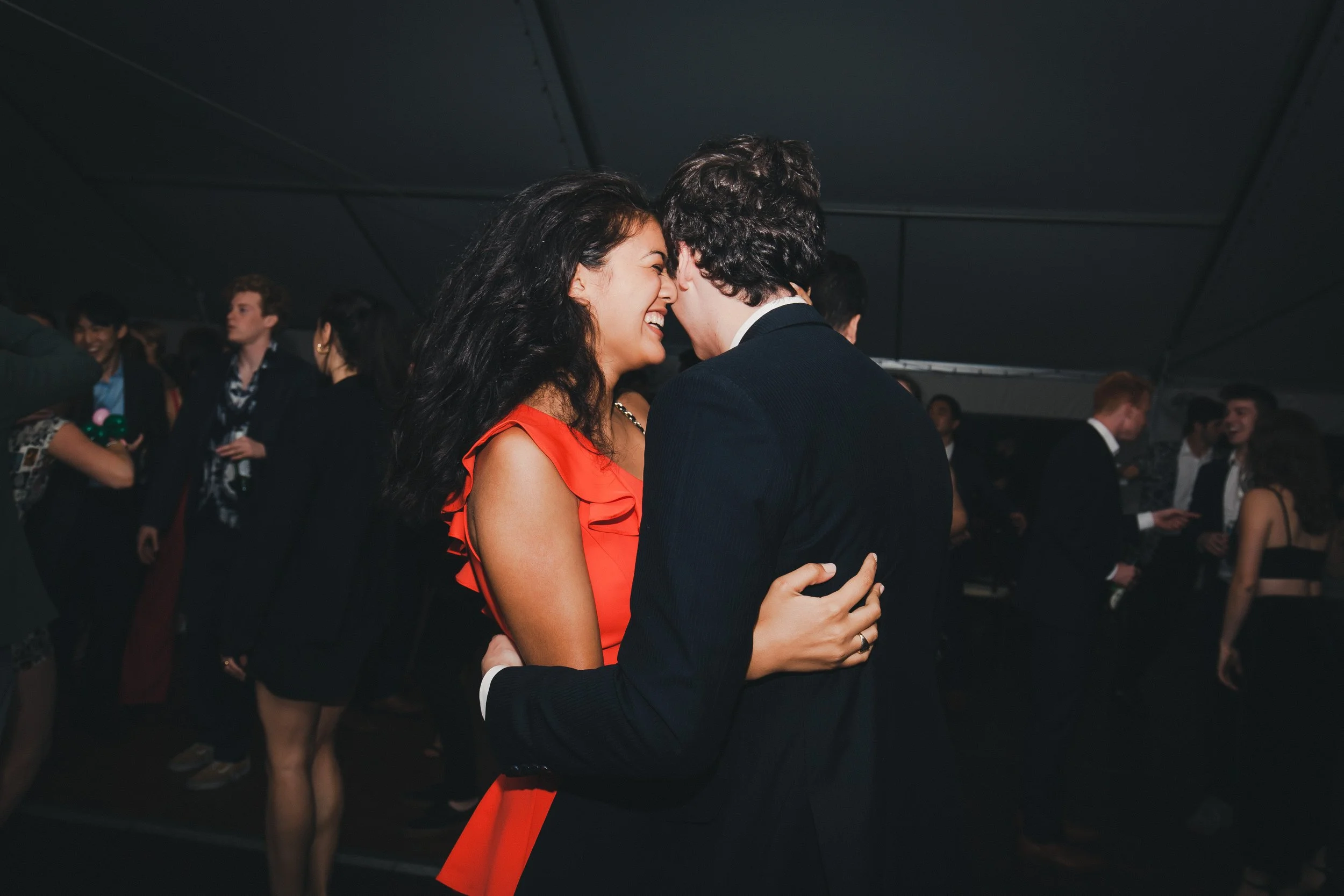 Couple dancing closely and smiling at a party or dance event, surrounded by other people in formal attire.