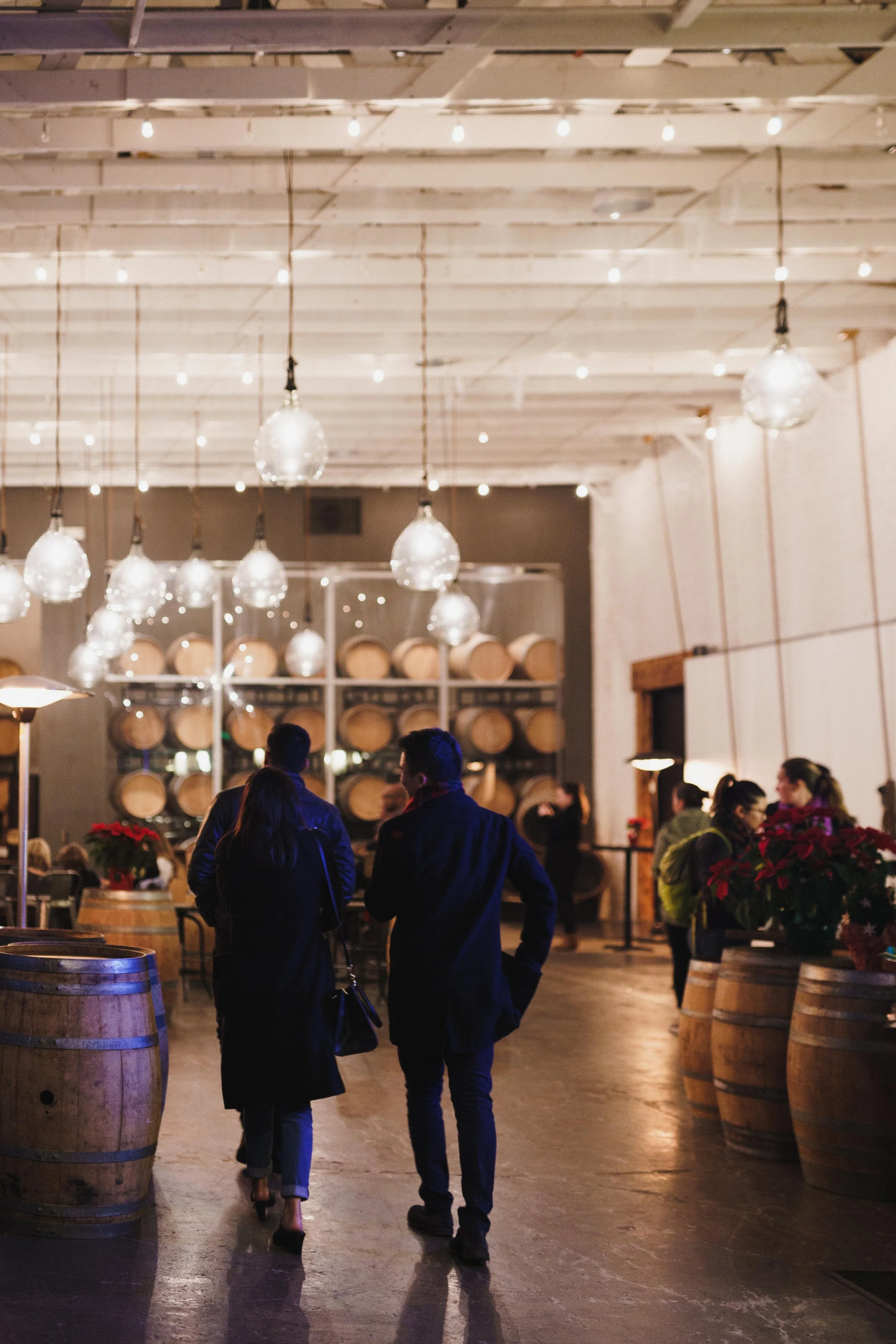 People walking inside a winery or wine cellar with barrels, hanging lights, and festive decor.