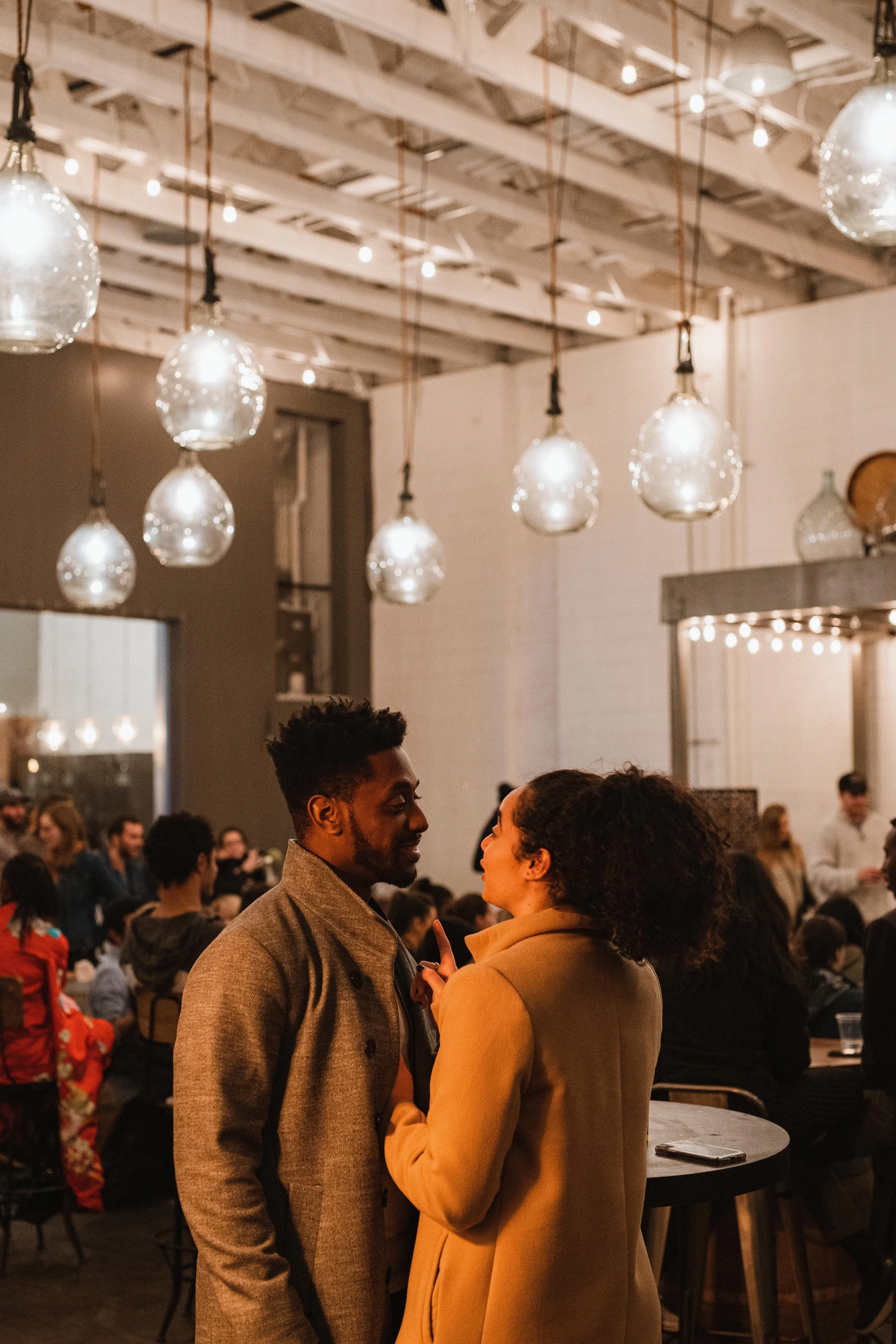 A man and woman having a conversation at a social gathering in a warmly lit venue with hanging glass pendant lights and many people seated at tables.