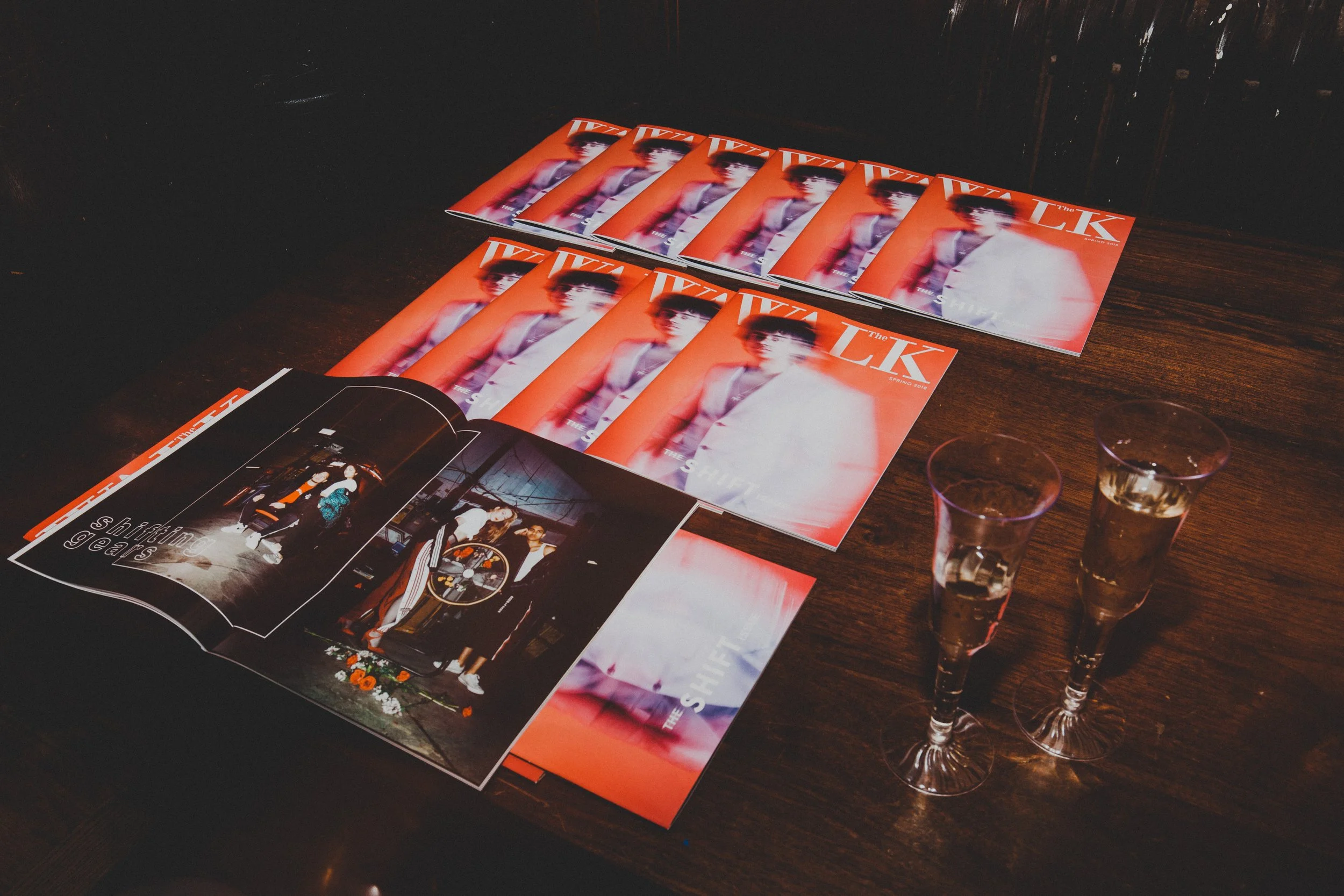 A wooden table holding several copies of a magazine with a portrait of a woman on the cover, a booklet open showing a group of people, and two glasses of champagne.