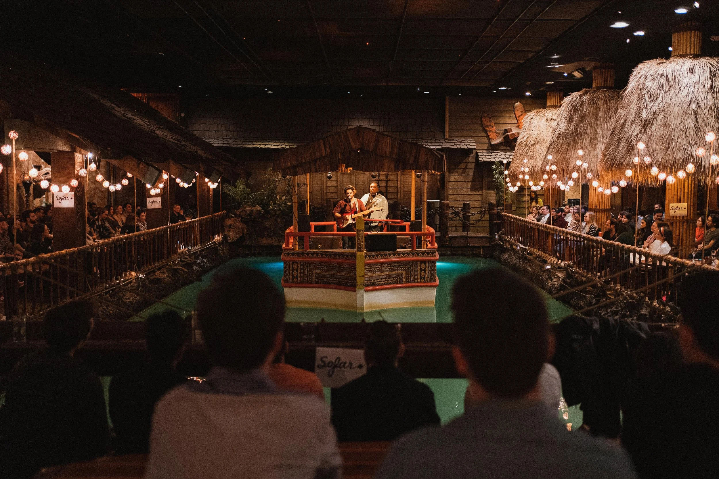 Indoor stage with two musicians performing, one playing guitar and the other singing, audience seated around a small pond, with traditional wooden and thatched hut decor, hanging string lights, and a tropical theme.