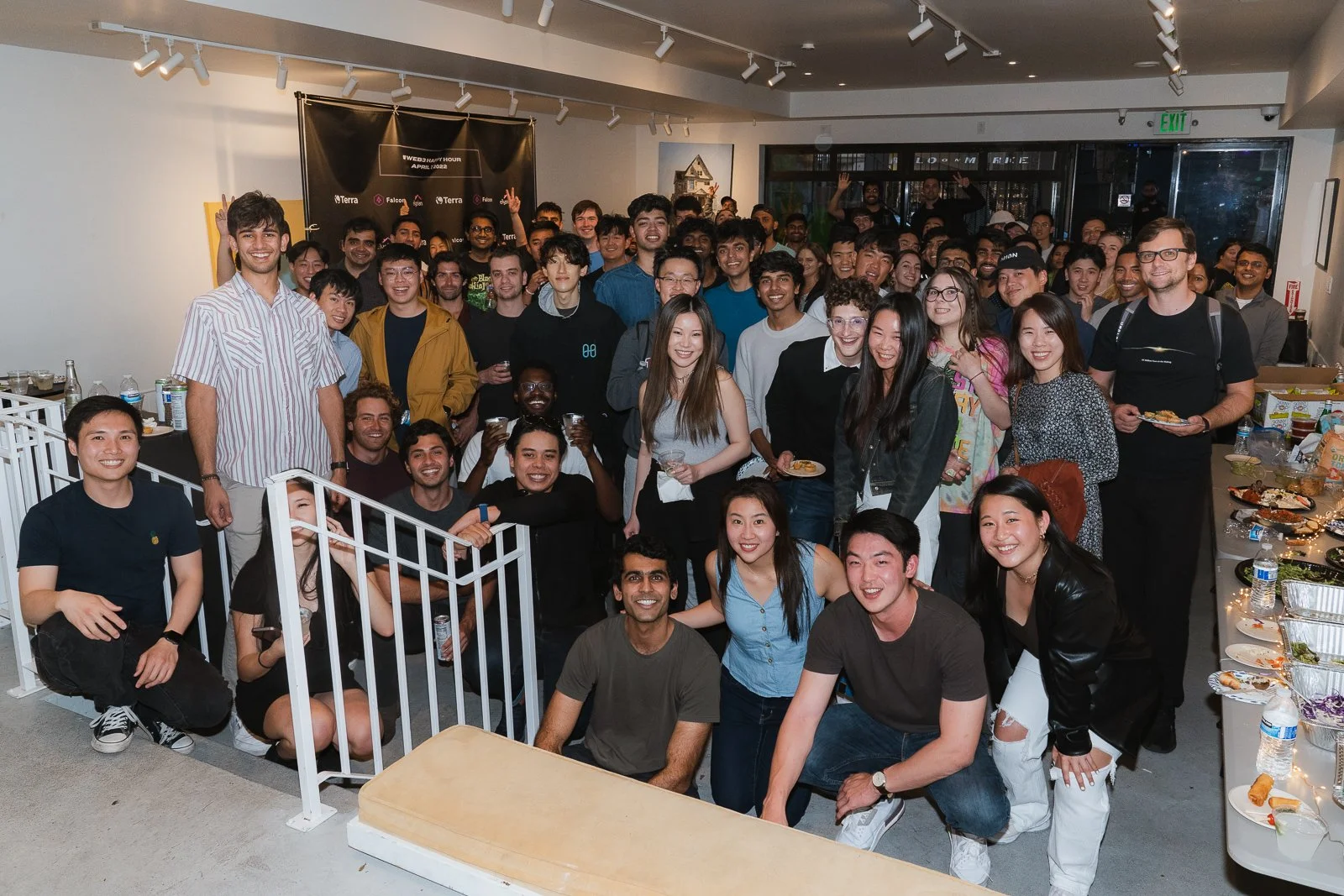Group of diverse young people gathered indoors at a celebration or event, smiling and posing for the photo with party snacks and drinks on tables nearby.