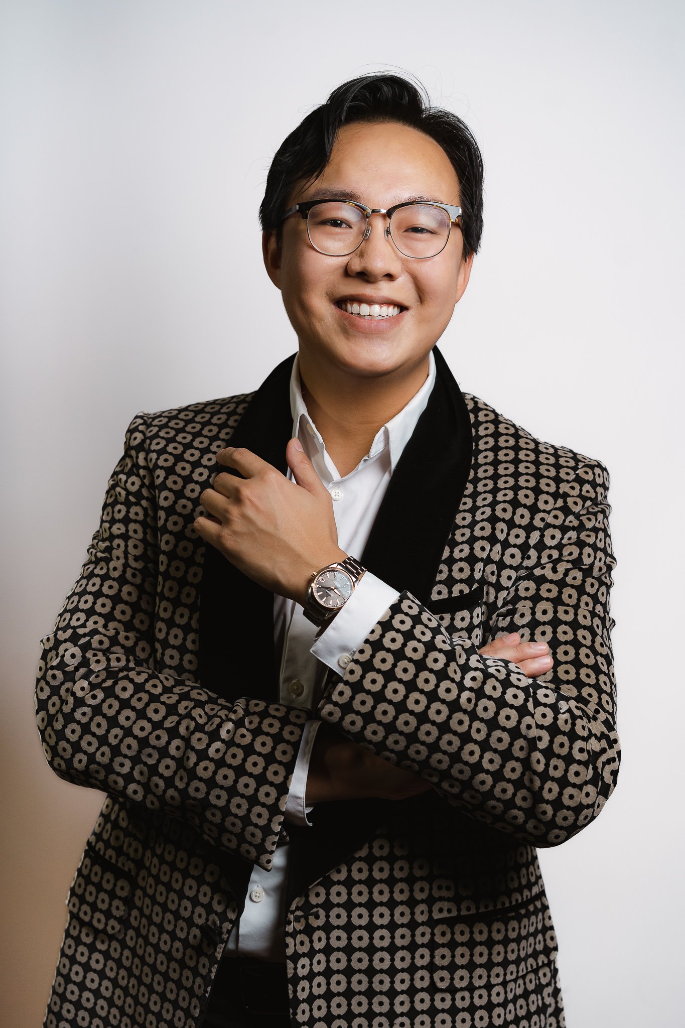 A young Asian man in a patterned blazer, white shirt, wearing glasses, smiling, with crossed arms and a wristwatch, standing against a plain background.