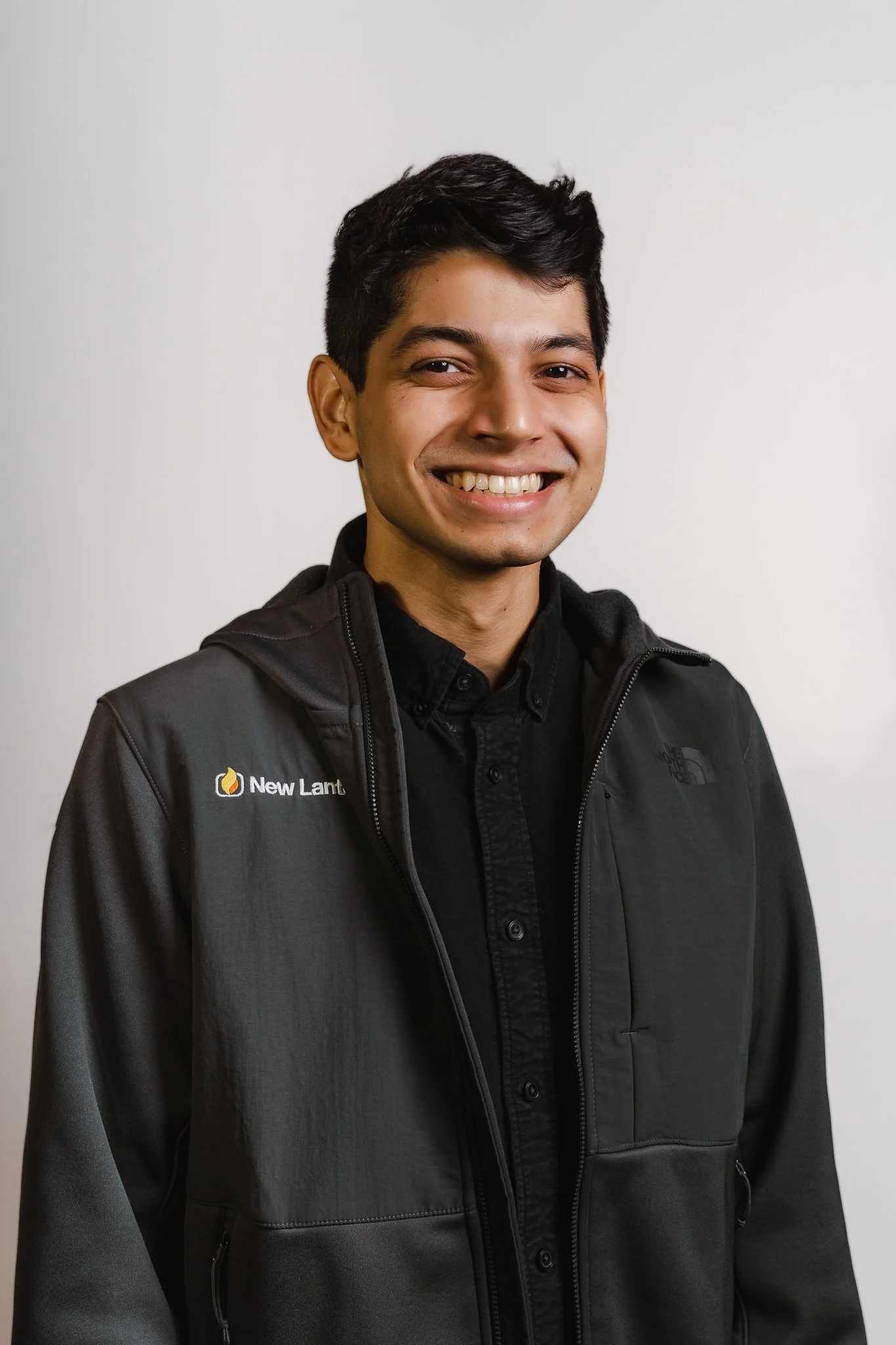 A young man with short black hair, smiling, wearing a black North Face jacket over a black shirt, standing against a plain white background.