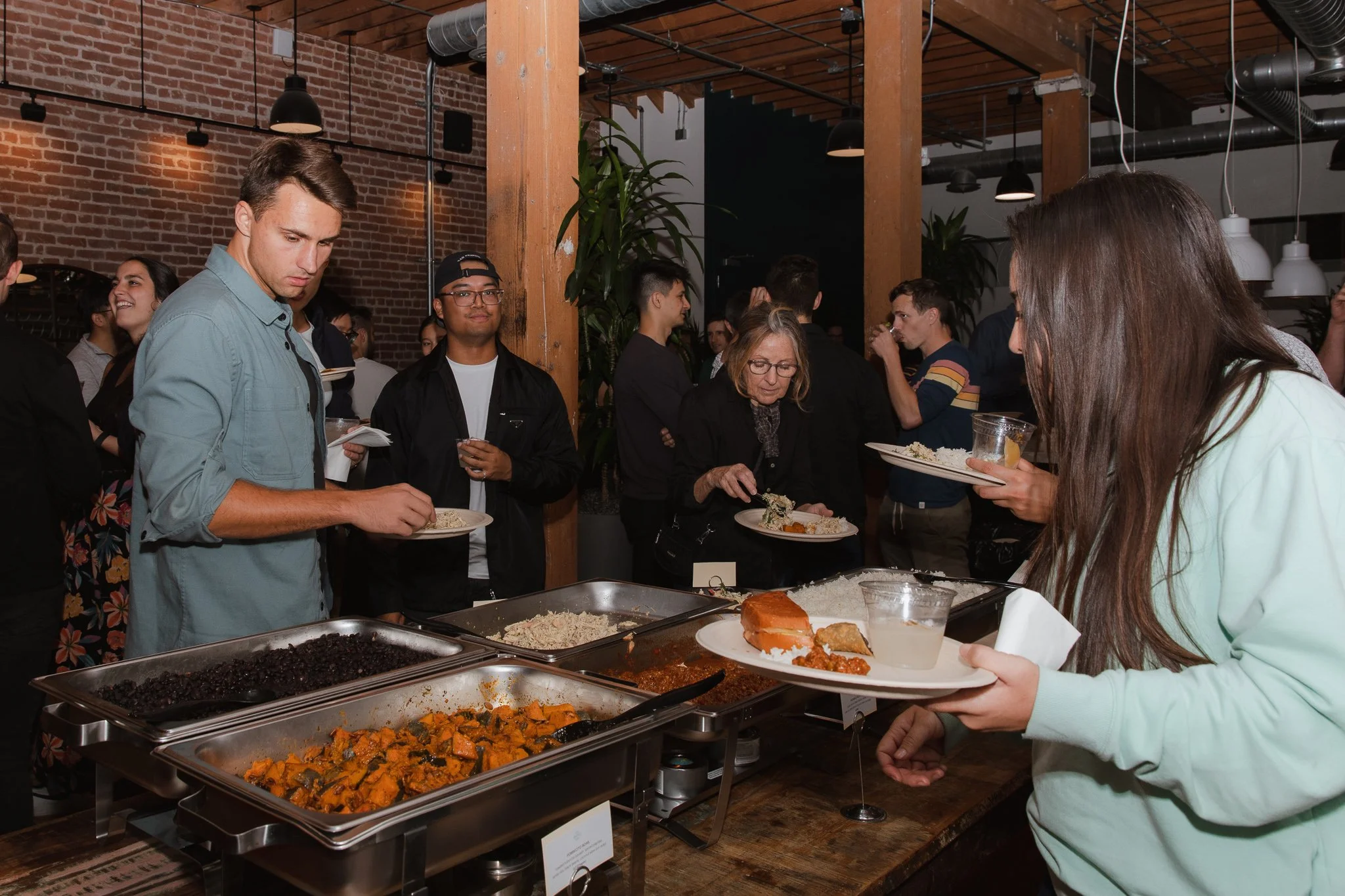 People lining up at a buffet with various dishes, including rice, vegetables, and meats, in a modern restaurant setting with brick walls and wooden beams.