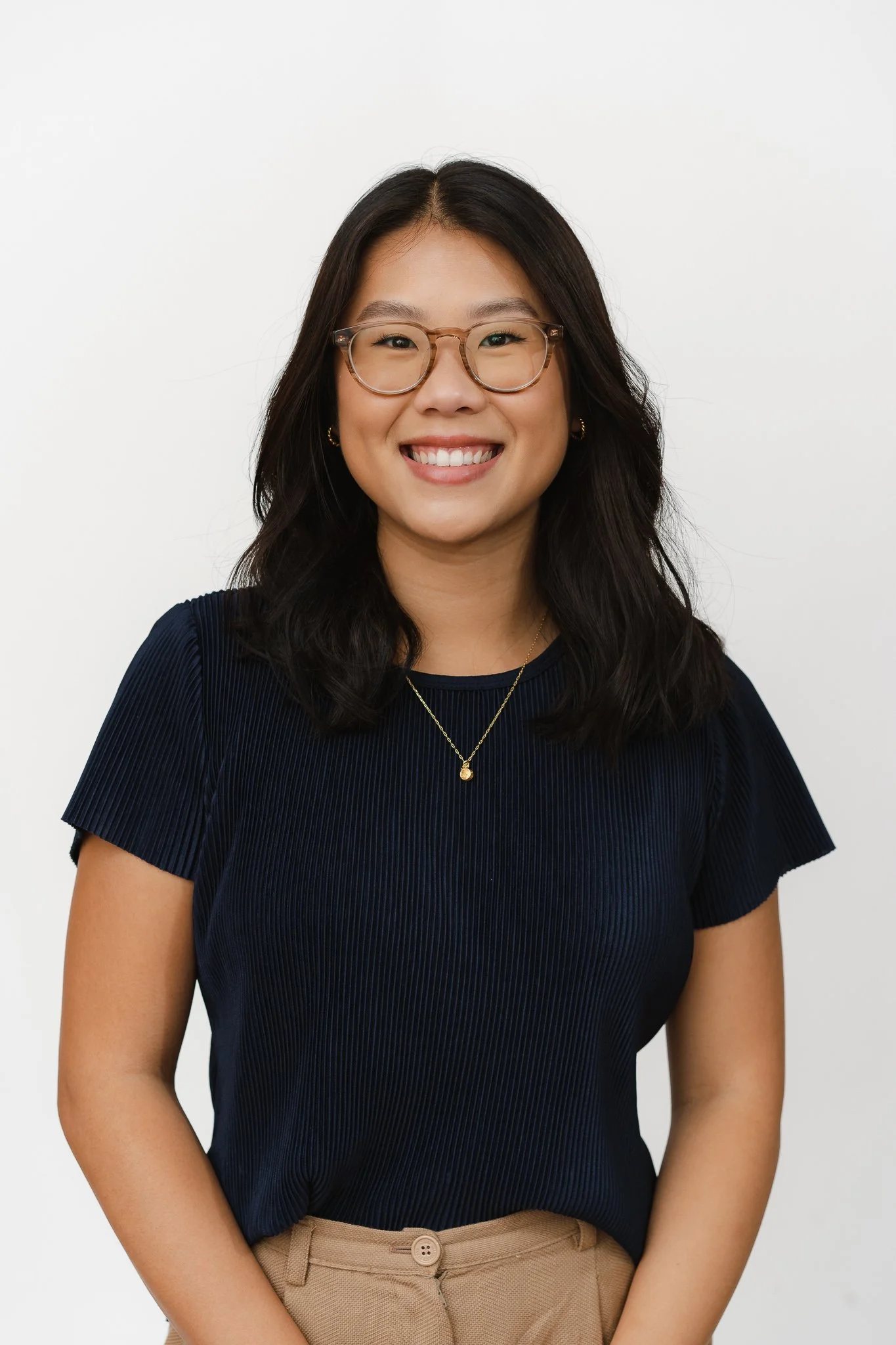 A young woman with shoulder-length dark hair, glasses, and a broad smile, wearing a navy blue top and beige pants, standing against a plain white background.