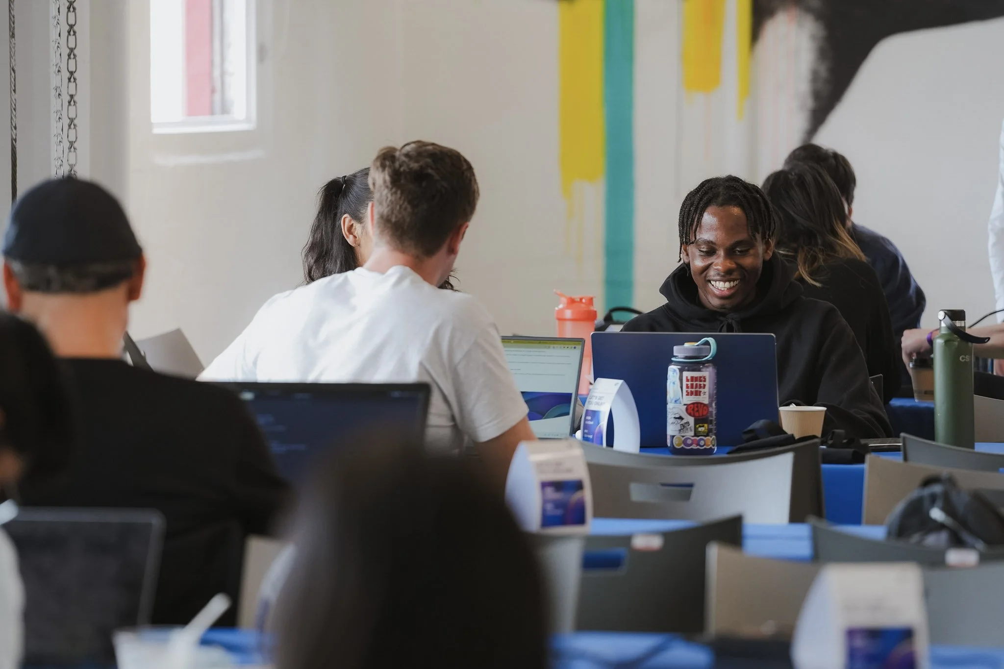 Group of young adults working on laptops in a brightly lit communal space, engaging and smiling.