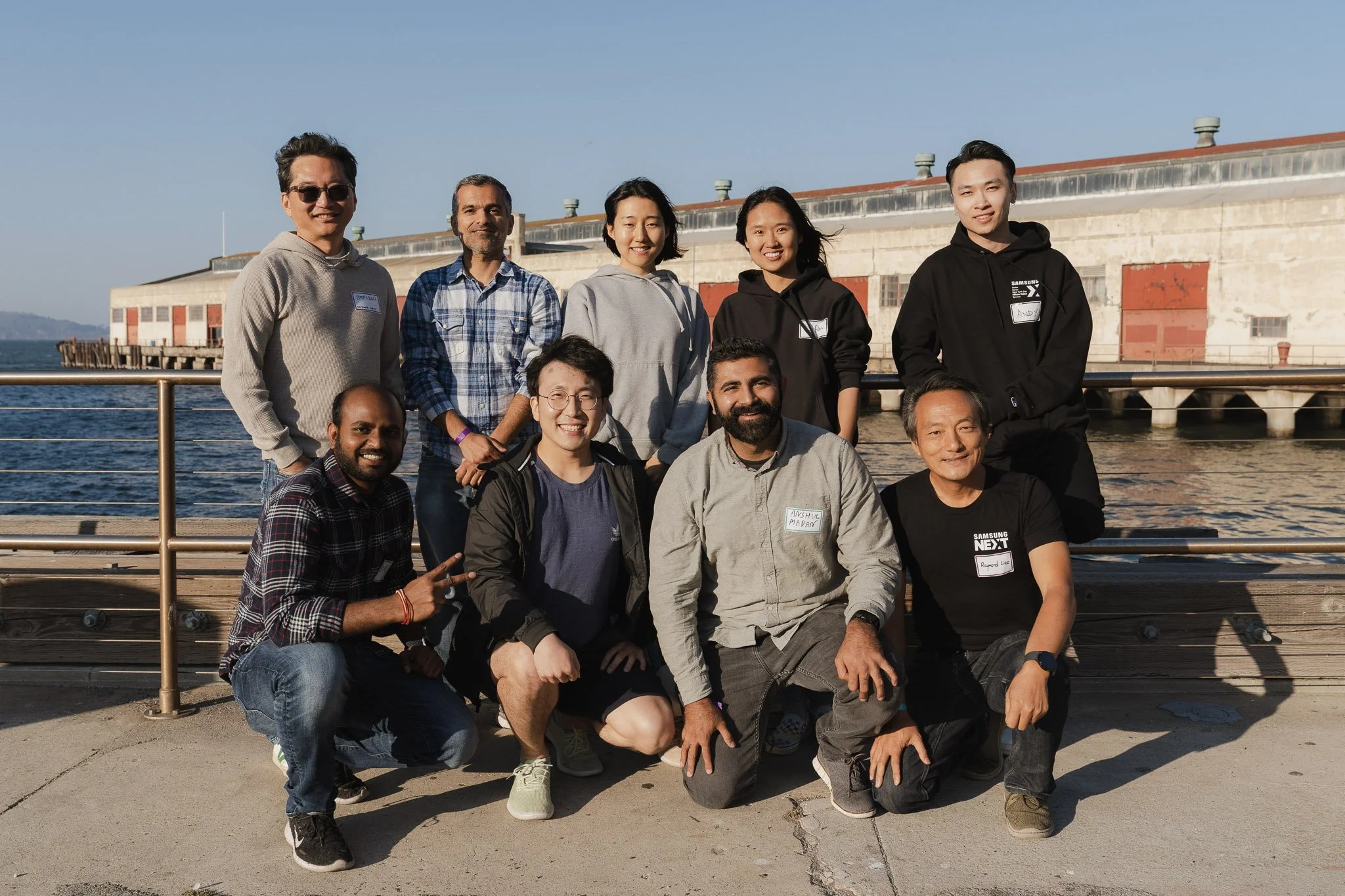 Group of ten diverse people posing outdoors near water with a building in the background.
