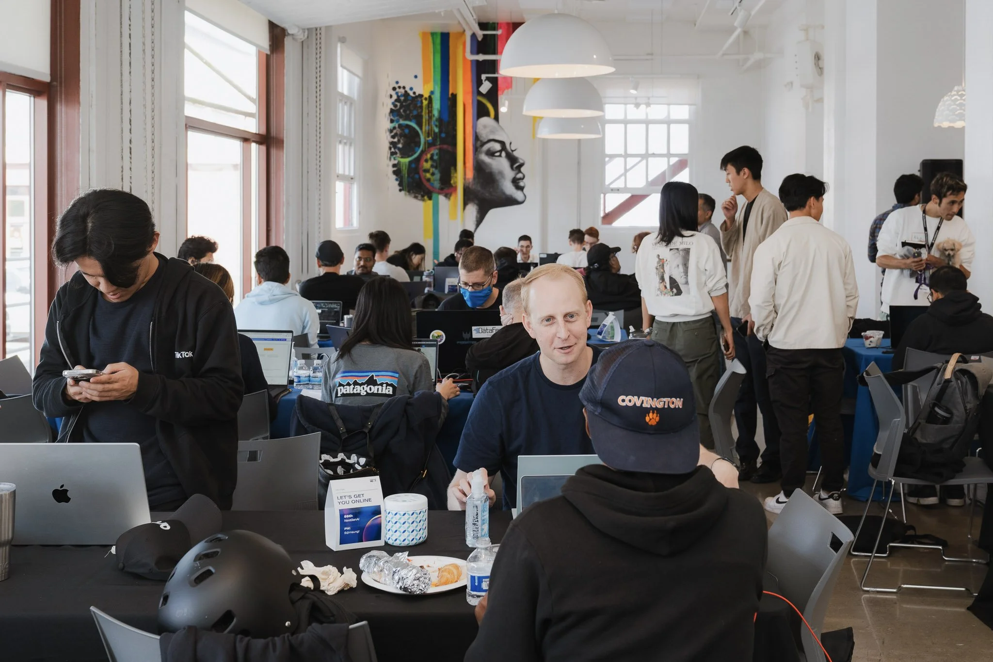 People working and socializing in a brightly lit, modern indoor space with group tables, laptops, and a large mural of a woman's face with colorful abstract art on the wall.
