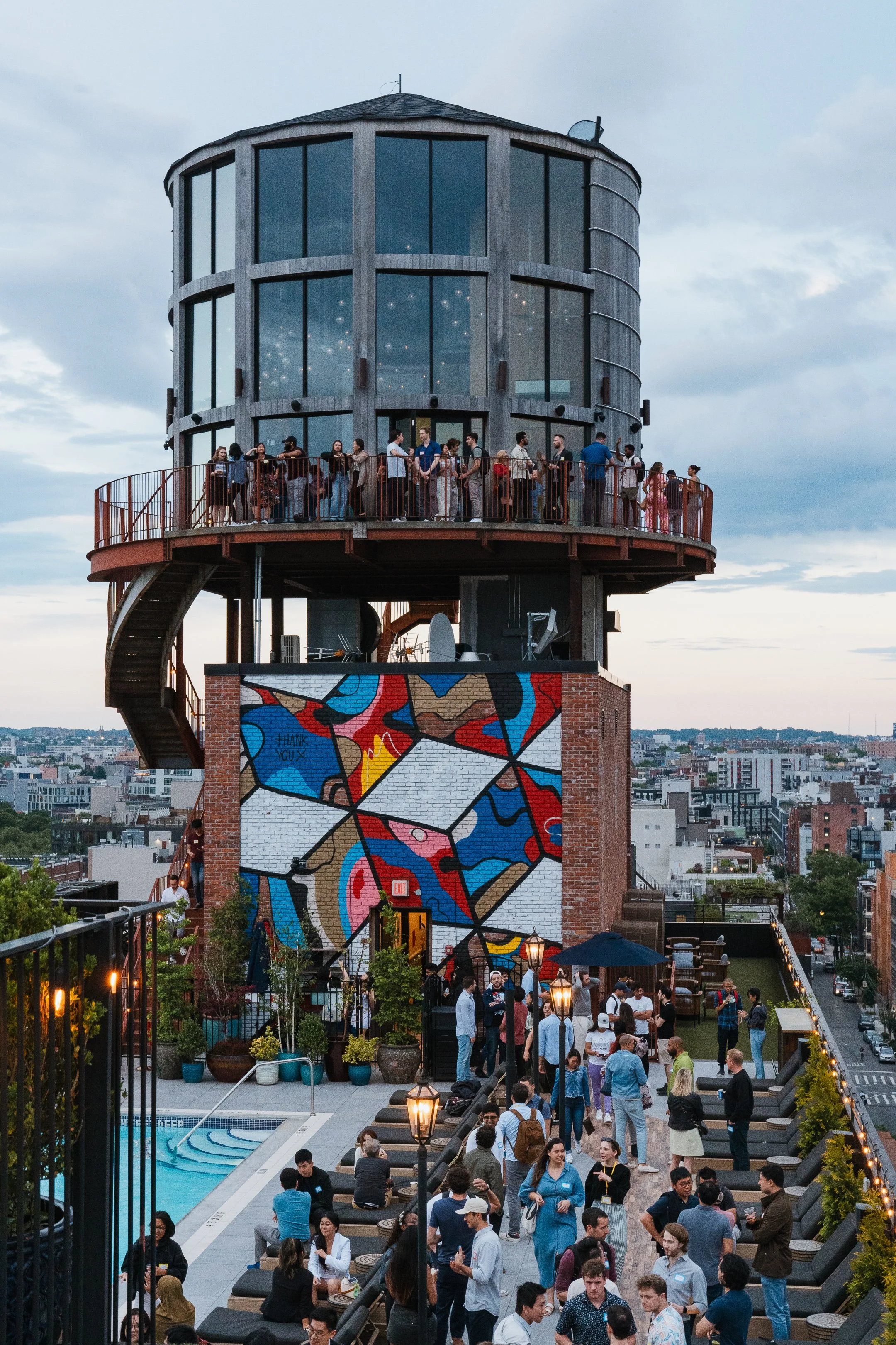 People gathering on a rooftop patio with a colorful mural wall, a pool, and cityscape in the background during evening.