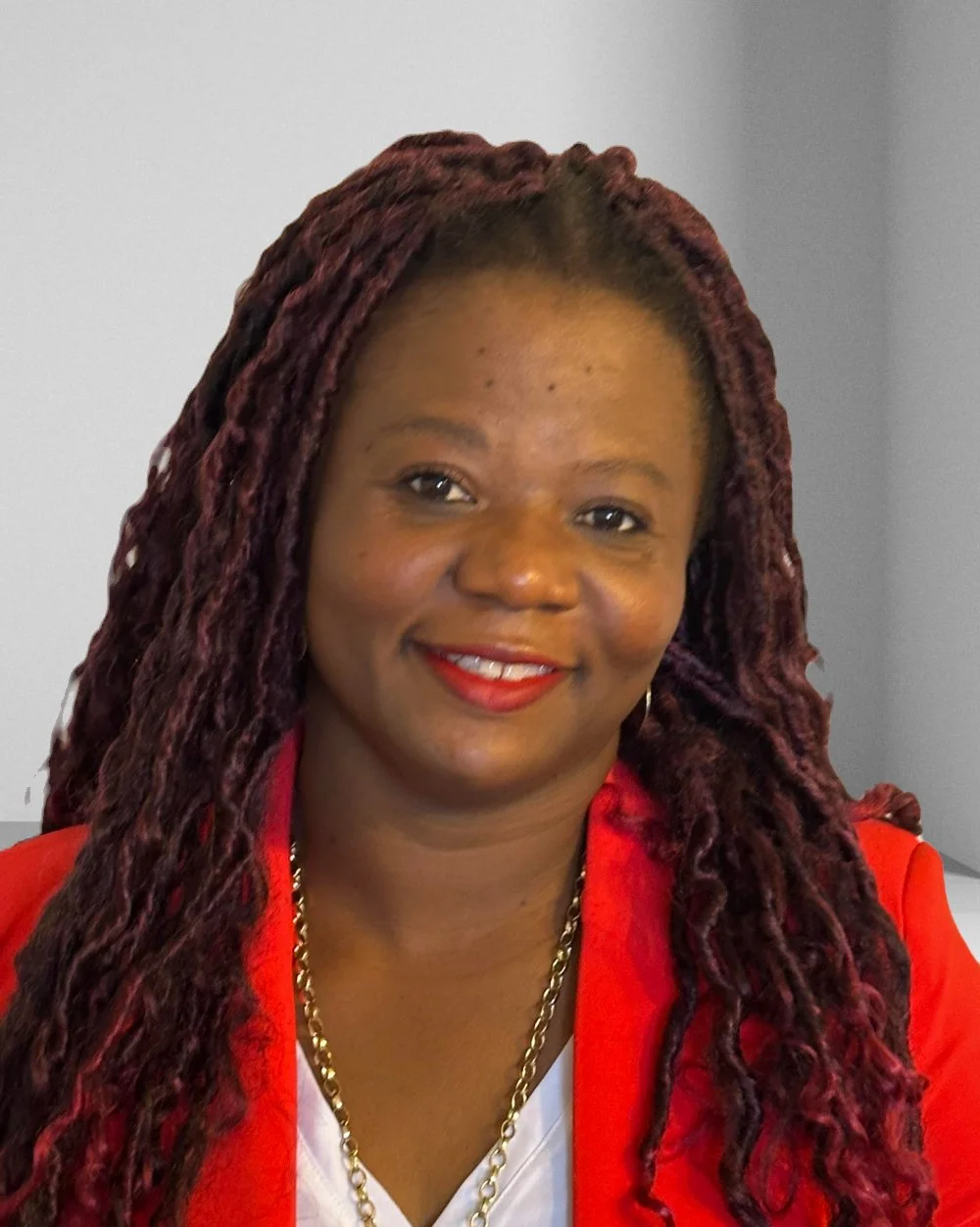 A woman with long red dreadlocks wearing a red blazer, white top, and gold chain necklace, smiling.