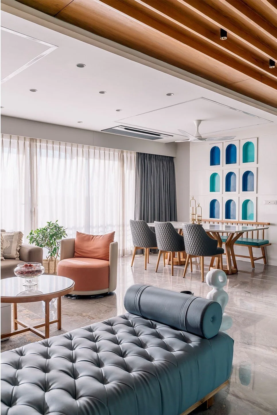 Modern living room with beige and pink armchair, grey tufted seating, round wooden coffee table, dining table with grey upholstered chairs, decorative wall niche with blue and teal accents, large window with sheer white curtains, and ceiling fan.