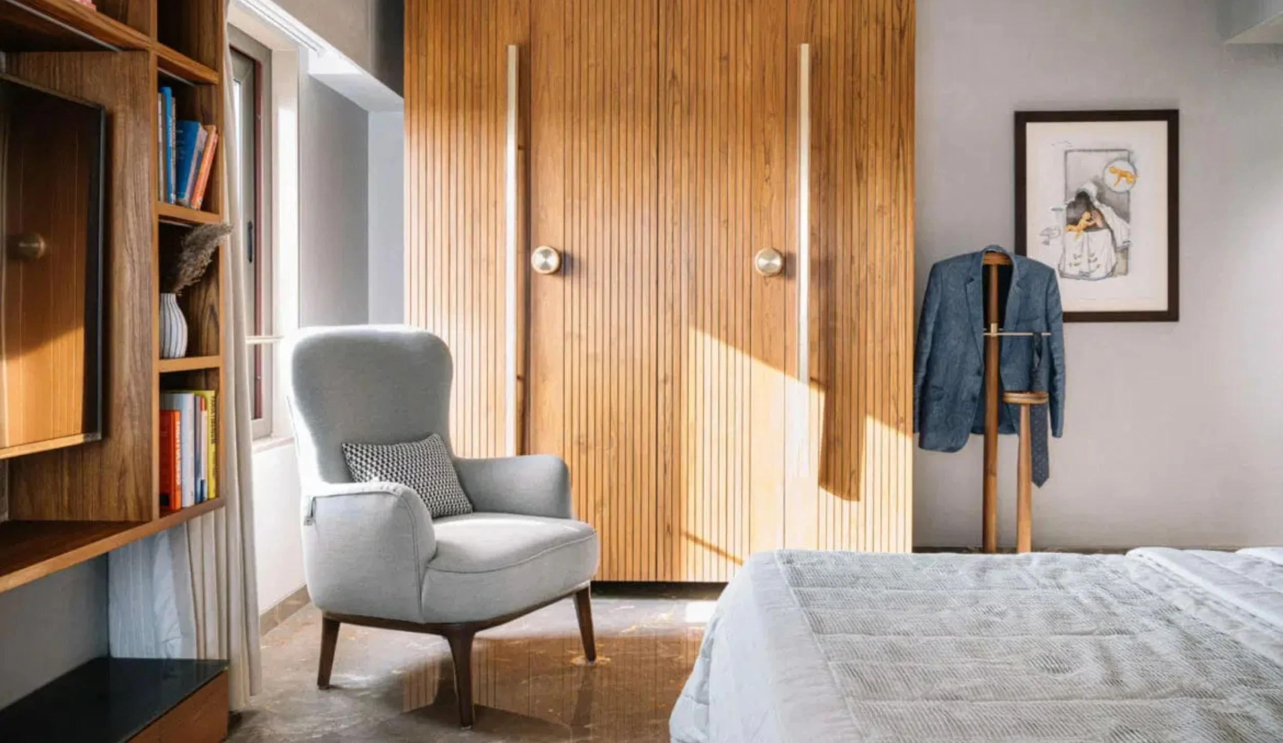 A bedroom with a white upholstered armchair, a gray pillow, wooden bookcase, wooden wardrobe, and a bed with light-colored bedding. There is a framed sketch on the wall, and a coat stand with a blue jacket hanging.