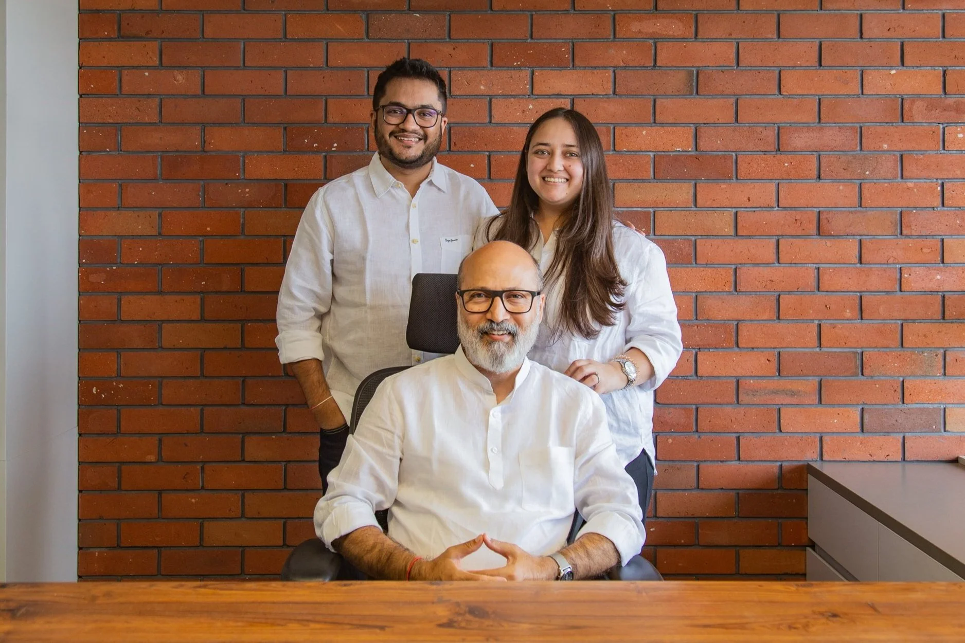 Three ethnically diverse professionals, two women and one man, posing together in an office with a brick wall in the background. The seated man has gray hair, glasses, and a beard, and is wearing a white shirt. The standing man has dark hair, glasses, and is also wearing a white shirt. The standing woman has long brown hair and is wearing a white shirt, smiling at the camera.