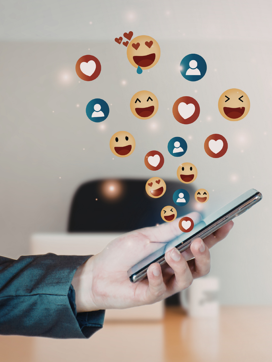 A person's hand holding a smartphone with floating emojis of hearts and smiling faces, indicating social media engagement.