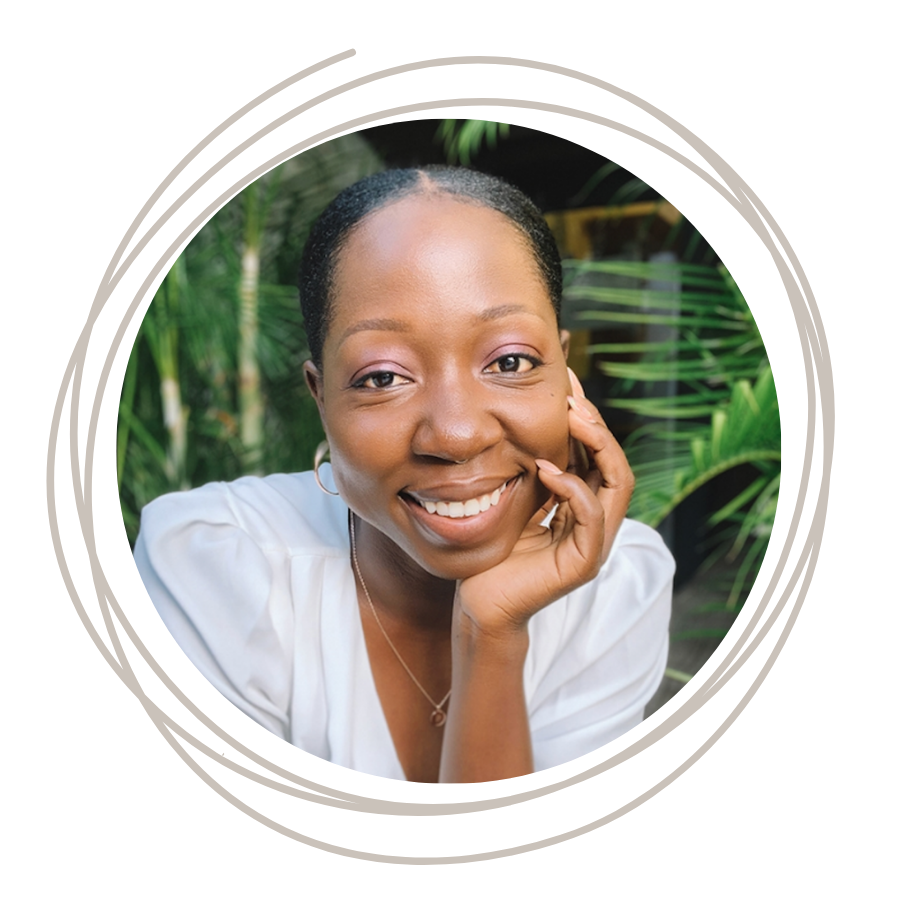 Close-up of a young smiling Black woman with natural hair, wearing a white top, outdoors with green foliage in the background.