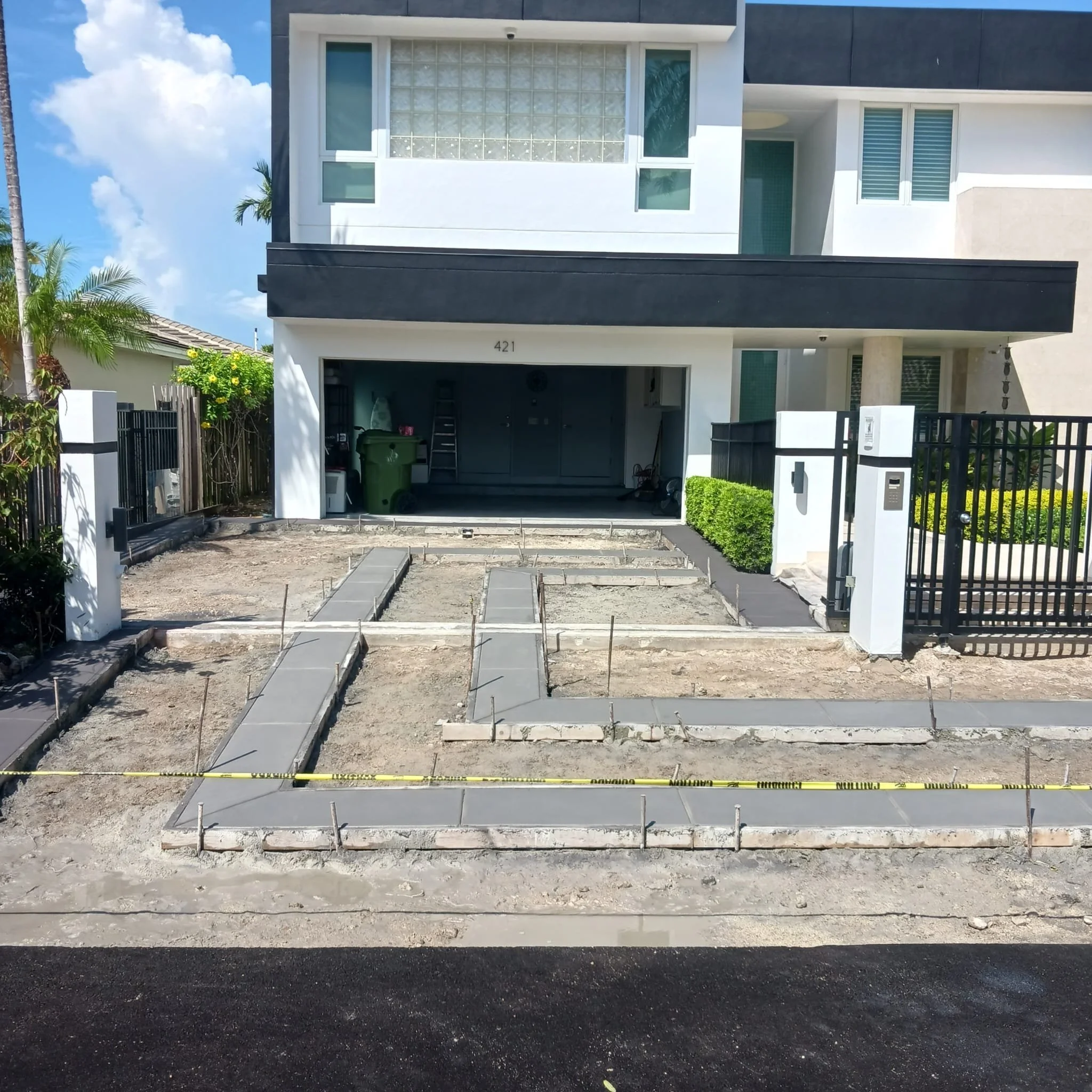 Front view of a modern two-story house with ongoing driveway construction, featuring newly laid concrete pathways and caution tape. The house includes a garage and modern design elements.