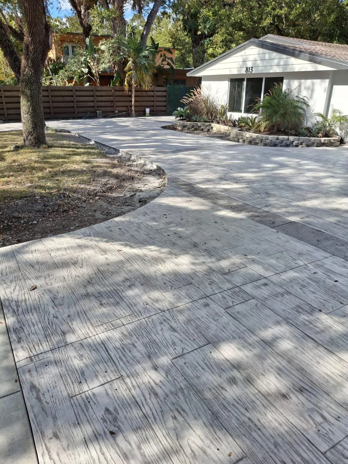 Curved concrete driveway leading to a house with a landscaped garden and palm trees, surrounded by a wooden fence.