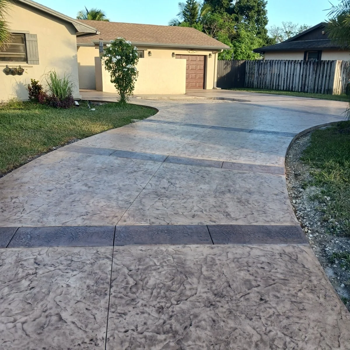 Concrete stamped driveway leading to a beige house with a brown garage door, surrounded by grass and a wooden fence in the backyard.