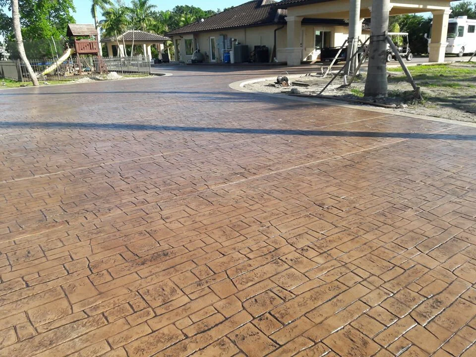 Stamped concrete driveway with brick pattern, surrounded by a house, trees, and a playground structure in the background.
