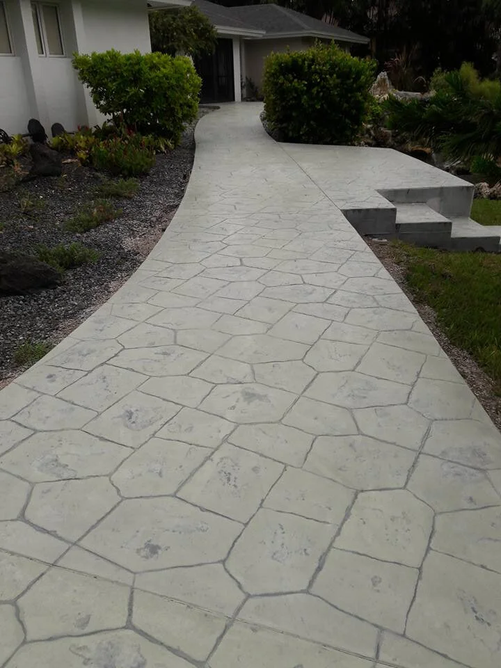 Decorative, colored, textured stamped concrete pathway with geometric pattern leading to a house, surrounded by greenery and shrubs.