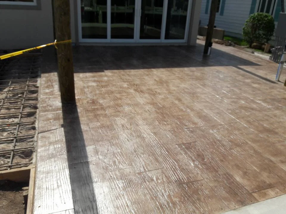 Stamped concrete patio with wood texture near a building with large windows, featuring a wooden post and caution tape.