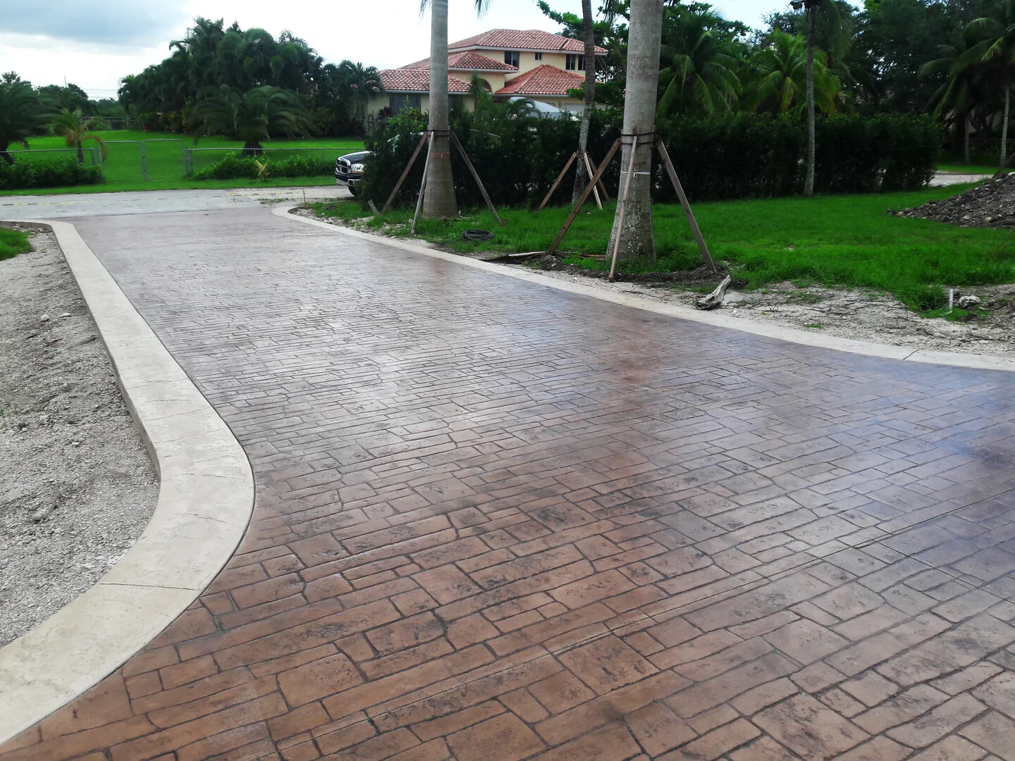 Stamped concrete driveway with brick pattern leading to a residential property with gardens and palm trees.
