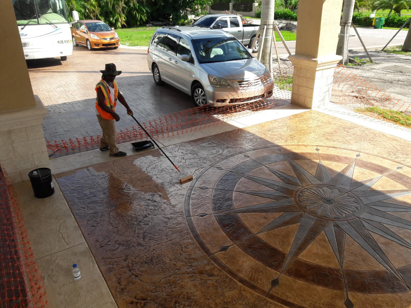 Worker applying sealant to decorative concrete floor with compass design outdoors, cars parked nearby.