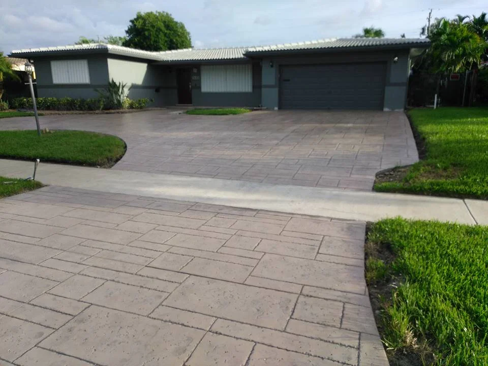 Concrete driveway leading to a single-story house with a garage, surrounded by green grass and landscaped plants, under a cloudy sky.