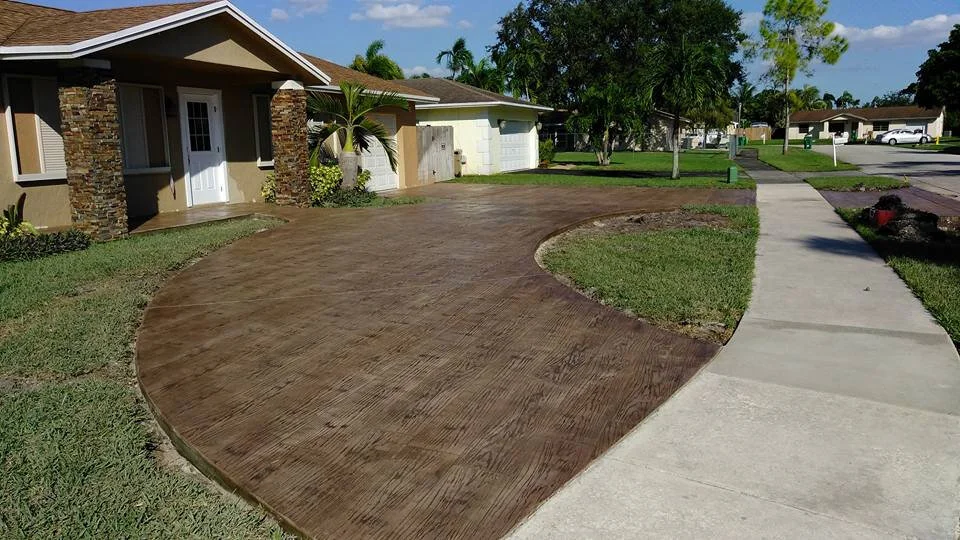 Curved decorative concrete driveway with textured surface leading to a suburban house with stone accents, next to a grassy lawn and sidewalk.