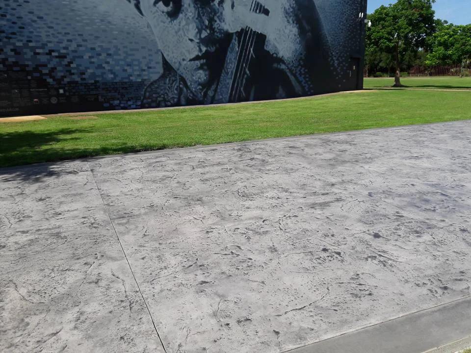 Mural of a person's face playing a string instrument on a wall, next to a grassy area and a smooth concrete path, surrounded by trees under a blue sky.