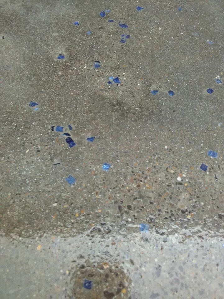 Close-up of a concrete surface with scattered small blue glass or tile fragments embedded in it, showing a speckled texture.