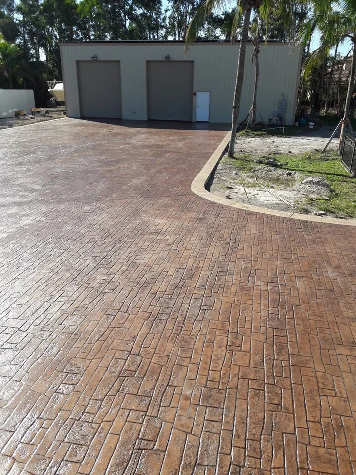 Large colored decorative stamped concrete driveway with brick pattern and separate color concrete edging leading to a garage with two large doors surrounded by trees.